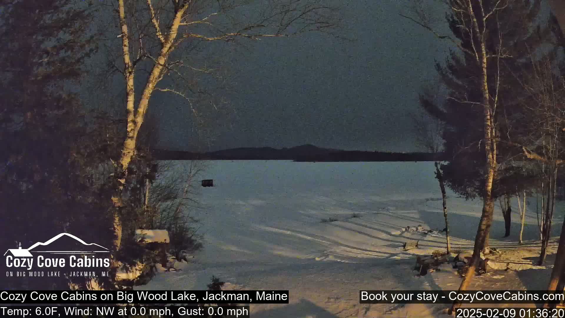 A partly frozen lake is seen from a snowy shoreline, with bare trees in the foreground under a dark, twilight sky.