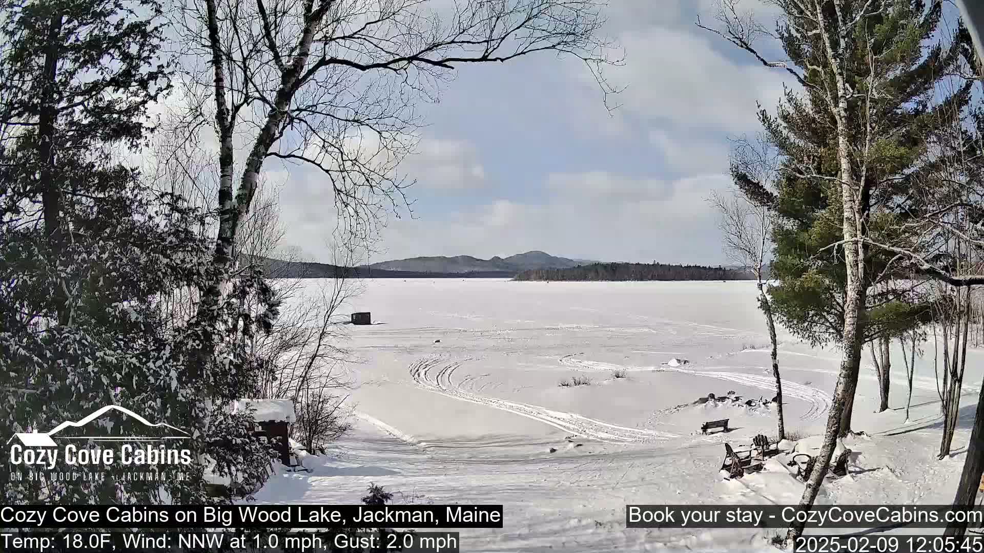 A snow-covered frozen lake with tire tracks and distant trees under a partly sunny sky.