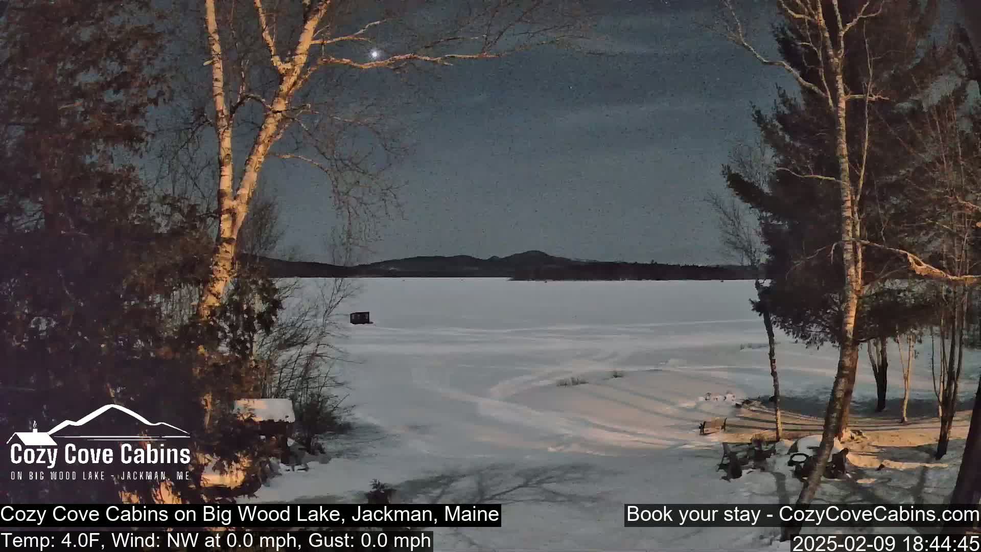 A snow-covered frozen lake is seen at twilight under a mostly clear sky, framed by bare winter trees.
