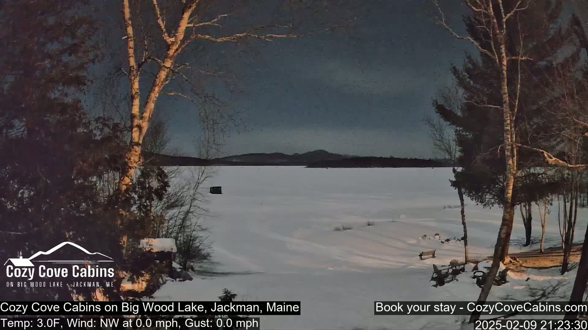 A snow-covered frozen lake is seen at dusk under a dark sky, with bare trees and a small structure in the foreground.