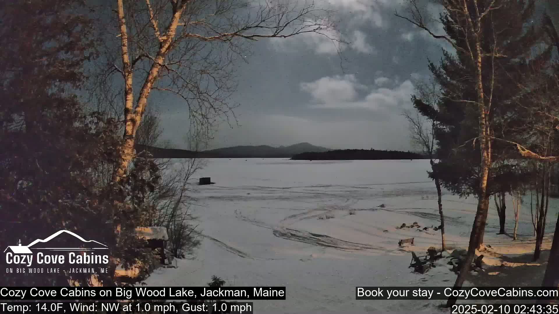 A snow-covered frozen lake is seen at twilight, under a partly cloudy sky, from a lakeside vantage point with bare trees in the foreground.