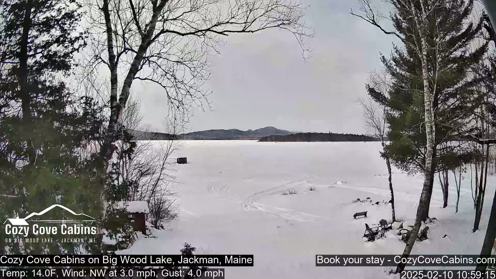 A snow-covered frozen lake with tire tracks and a few small structures is seen from a snowy lakeside vantage point, under an overcast sky with a light wind.