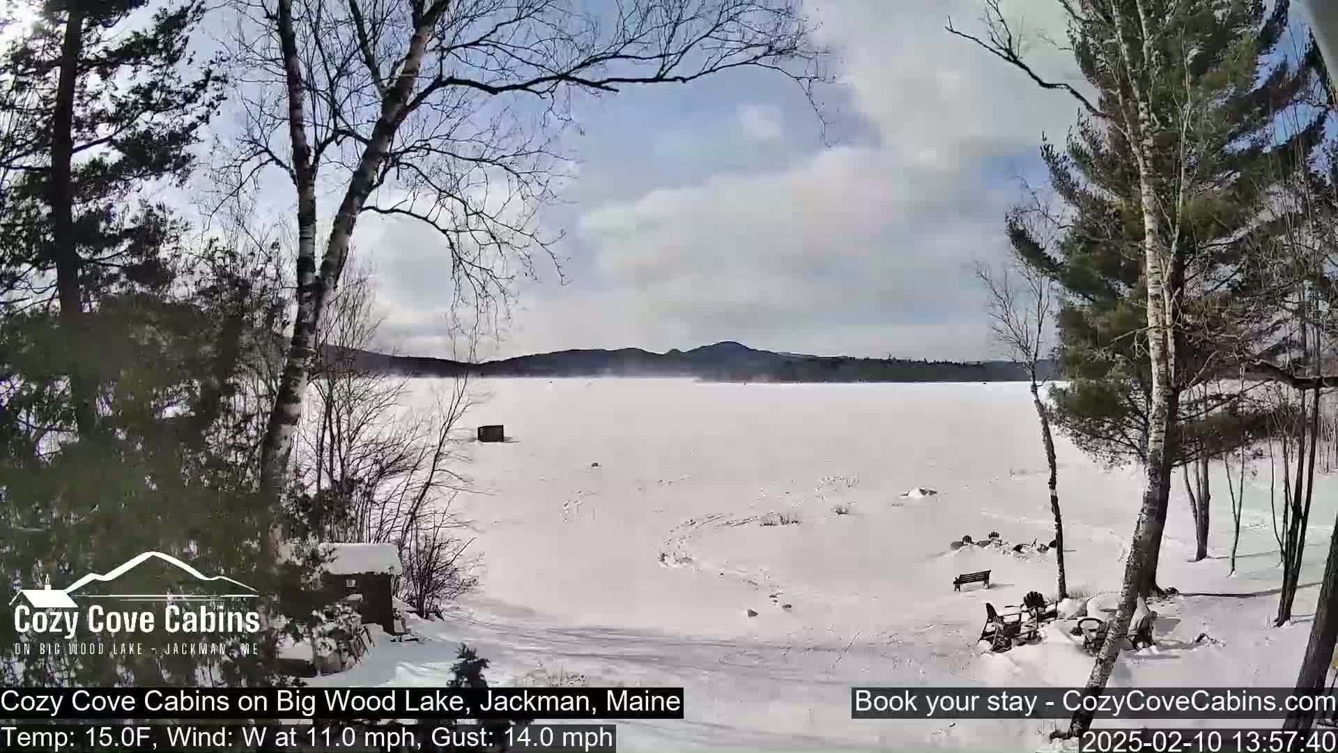 A snow-covered frozen lake is visible from a snowy shoreline, with leafless trees and a cabin in the foreground under a partly cloudy sky; the wind is blowing from the west.