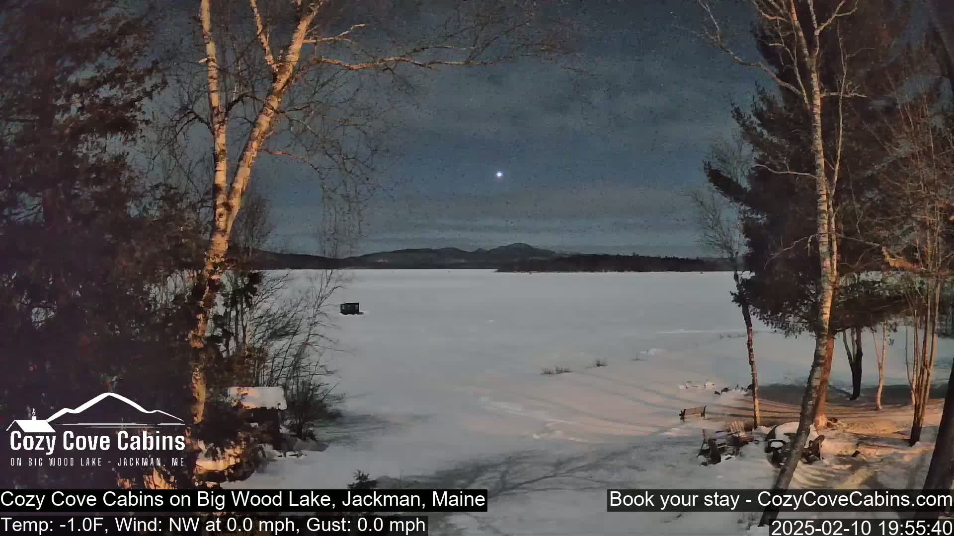 A snow-covered lake is seen at twilight under a mostly clear sky, with bare trees in the foreground and distant mountains in the background.