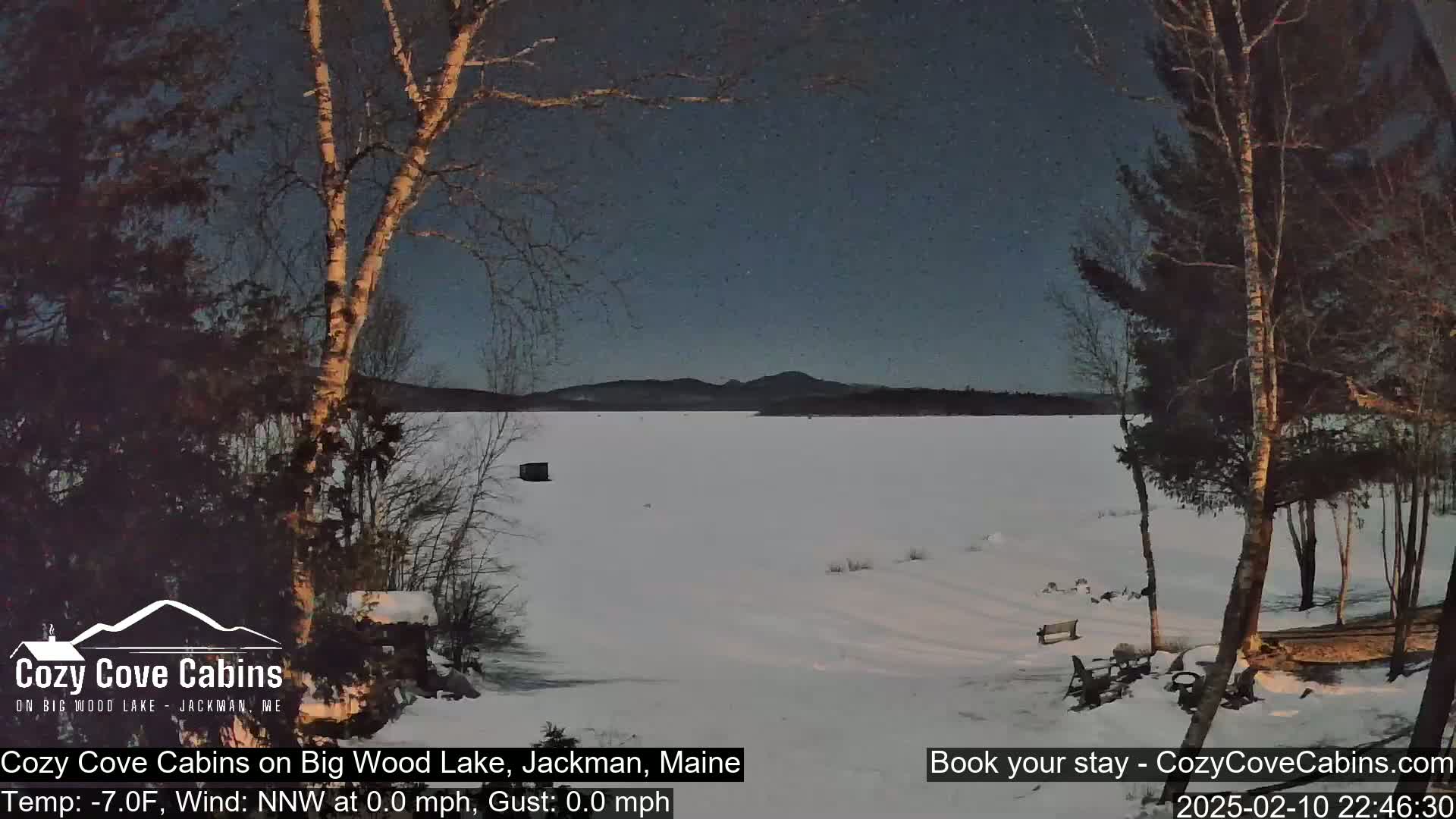 A snow-covered frozen lake is seen at dusk, under a dark blue, clear sky, with bare trees and a small structure visible in the foreground.