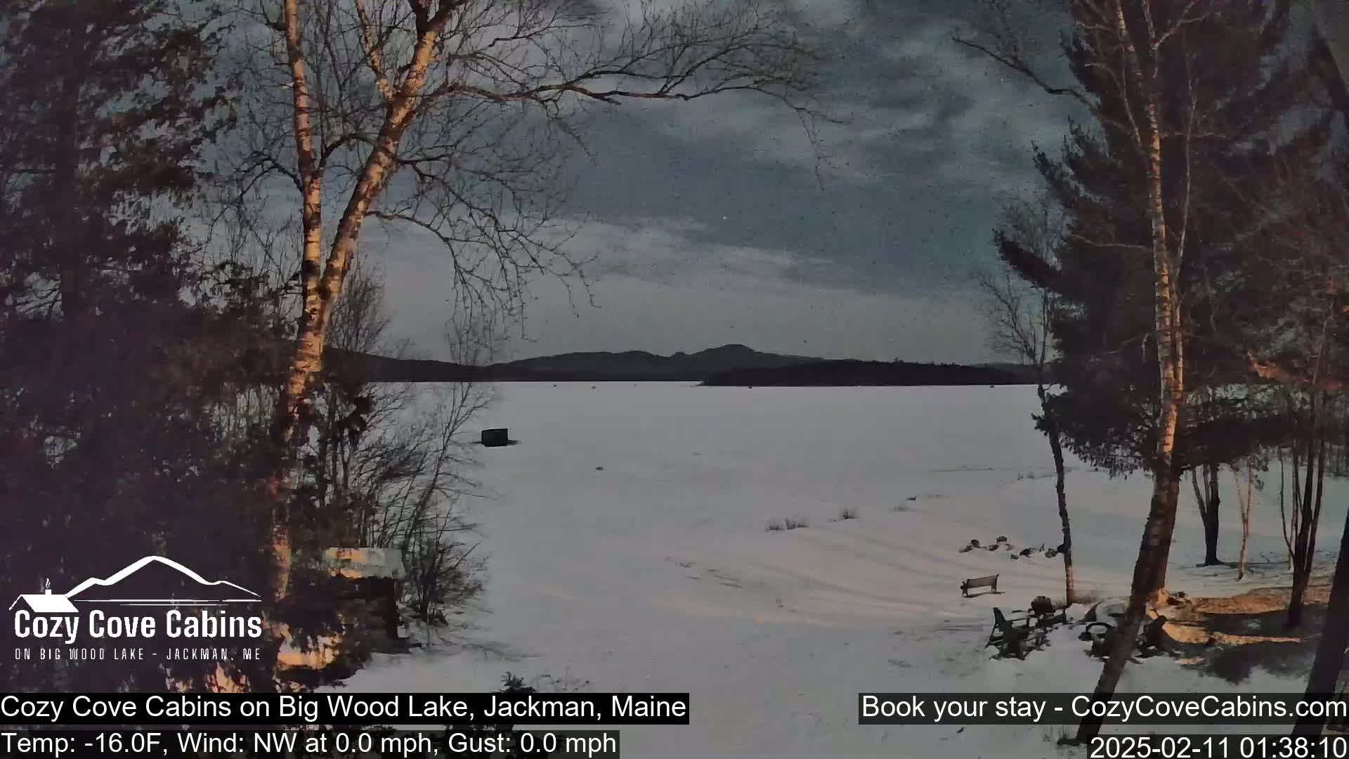A snow-covered frozen lake is viewed from a snowy shoreline, under a dark, cloudy sky, with bare trees and a small structure visible in the distance.