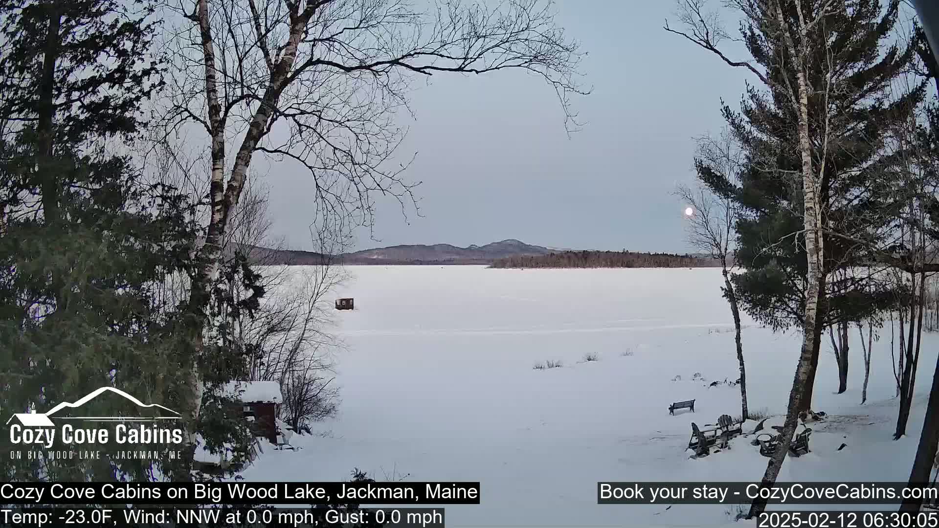 A snow-covered frozen lake with a distant mountain range is viewed from a snowy lakeside, under a pale sky with a bright moon.