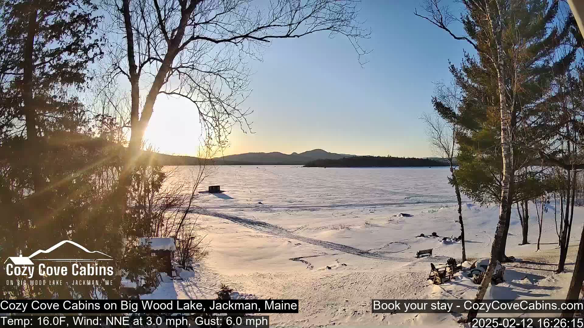 A snow-covered frozen lake at sunset, viewed from a snowy shoreline with trees in the foreground and distant mountains.