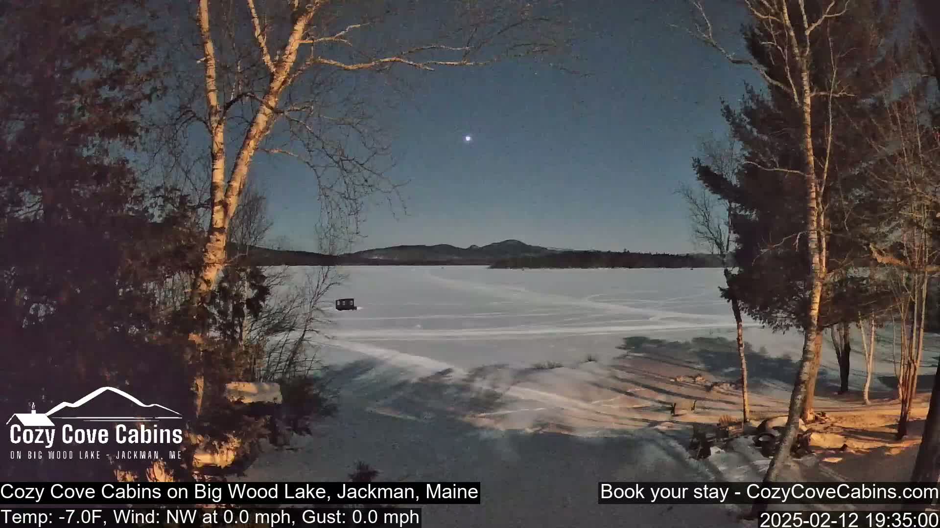A frozen lake is visible under a twilight sky, with bare trees in the foreground and a distant mountain range.