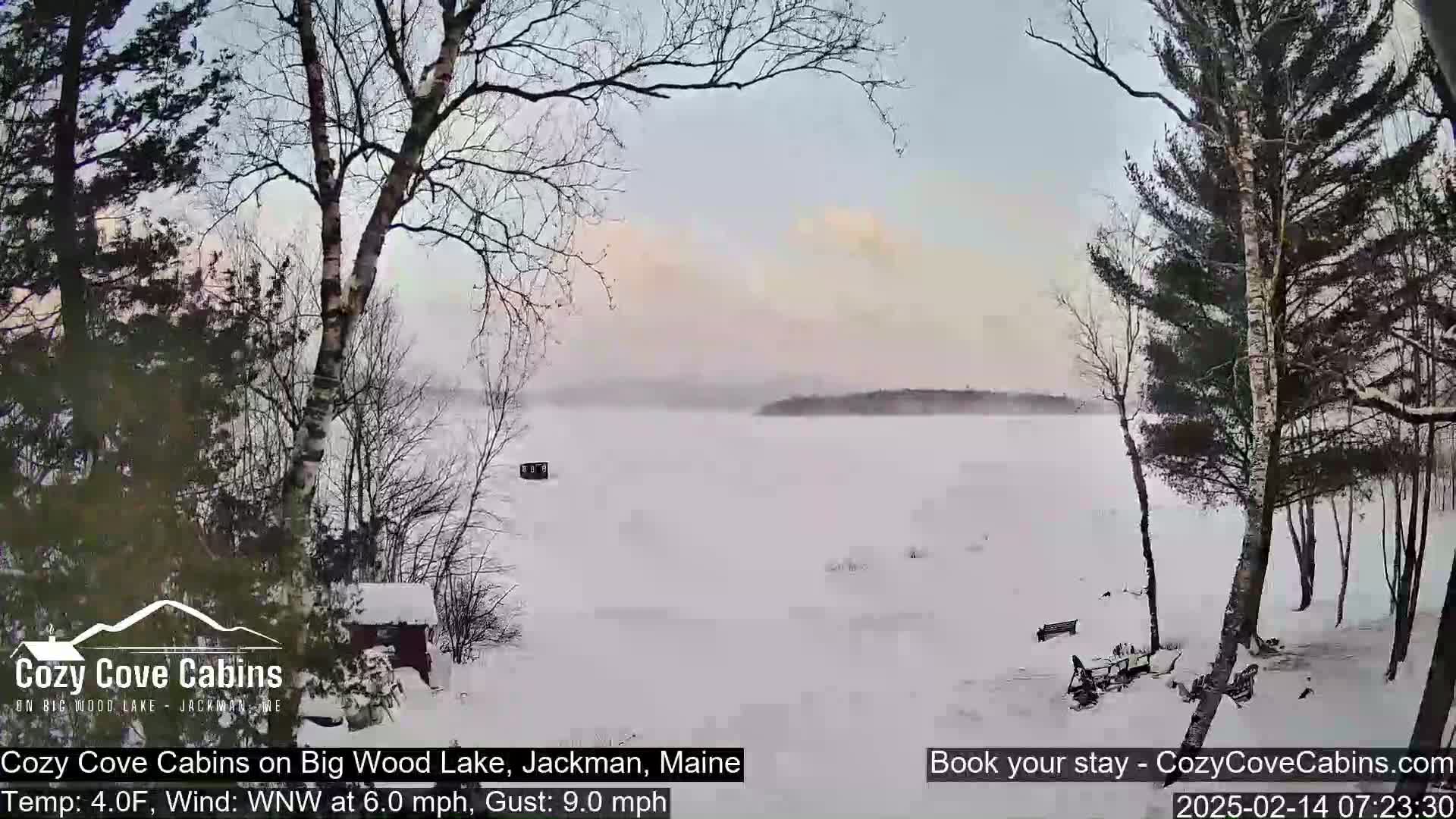 A snow-covered lake is seen from a snowy, wooded shoreline under a hazy, light sky with a wind of 6.0 mph and gusts up to 9.0 mph.