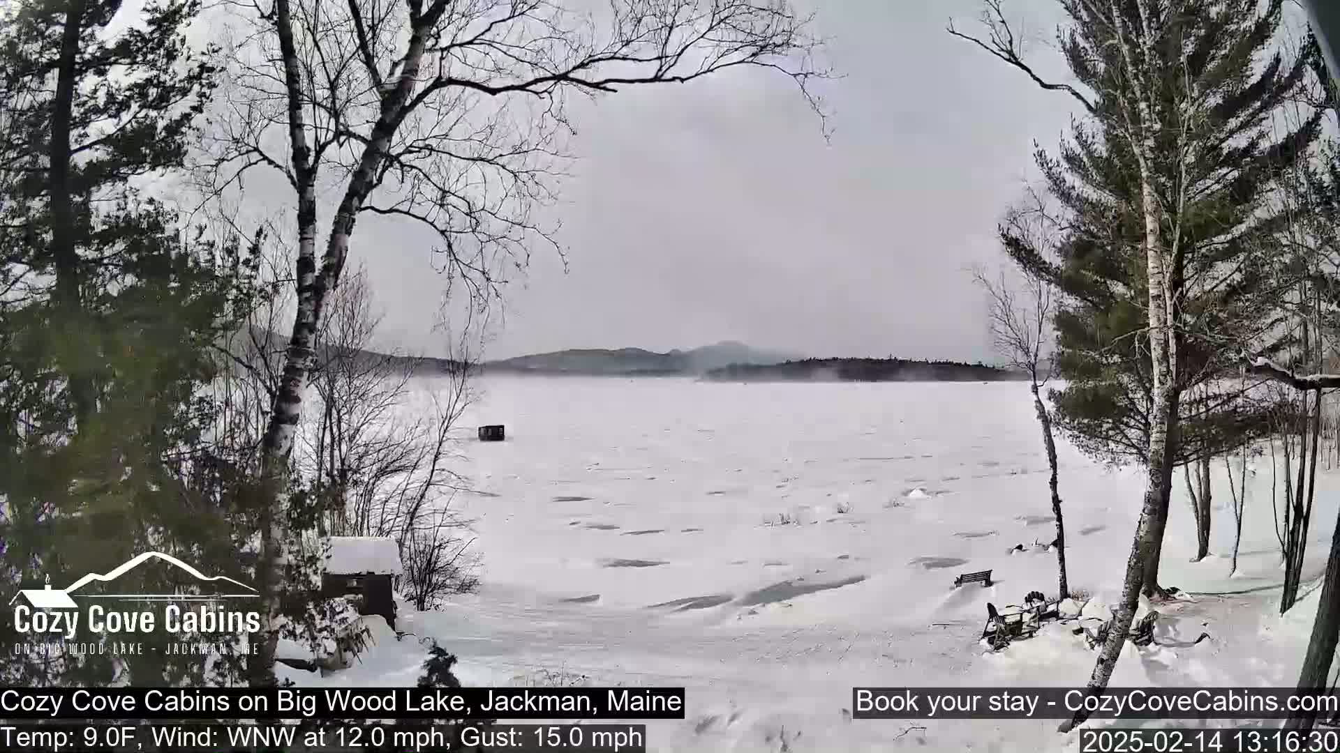 A snow-covered, partially frozen lake is viewed from a snowy shoreline with leafless trees and a small structure under an overcast sky.