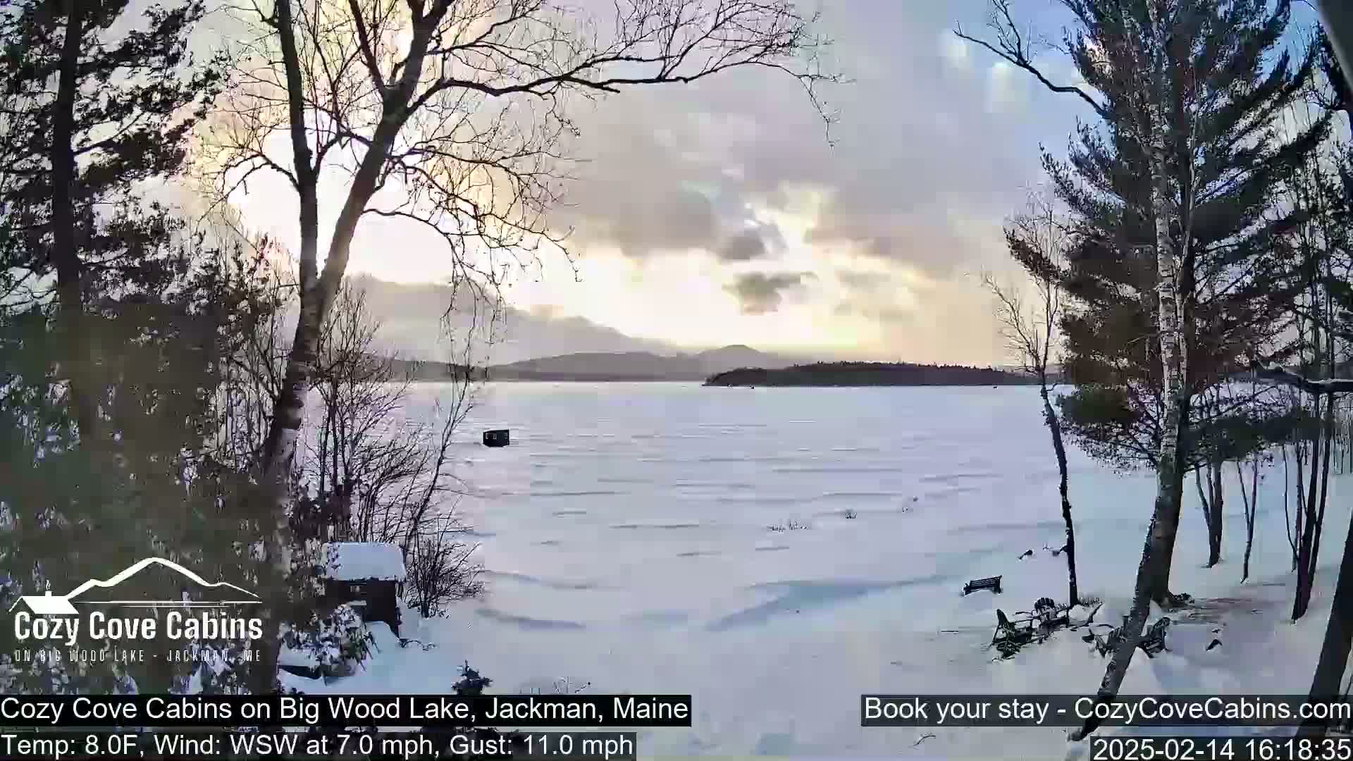 A snow-covered frozen lake with distant mountains under a partly cloudy sky, viewed from a snowy shoreline with leafless trees.