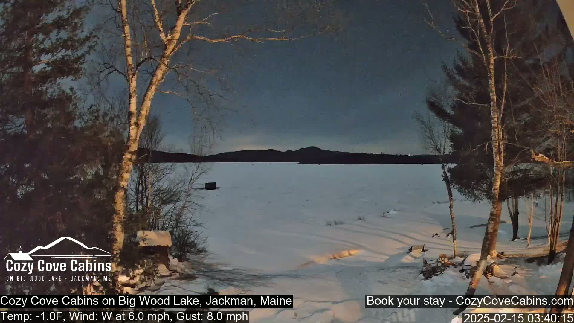 A snow-covered frozen lake is seen at night under a twilight sky, with bare trees in the foreground and distant mountains in the background, in cold, windy conditions.