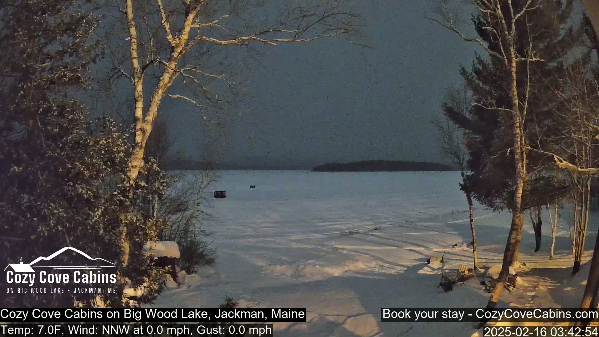 A snow-covered, frozen lake is visible from a snowy shoreline with leafless trees under a dark, twilight sky.