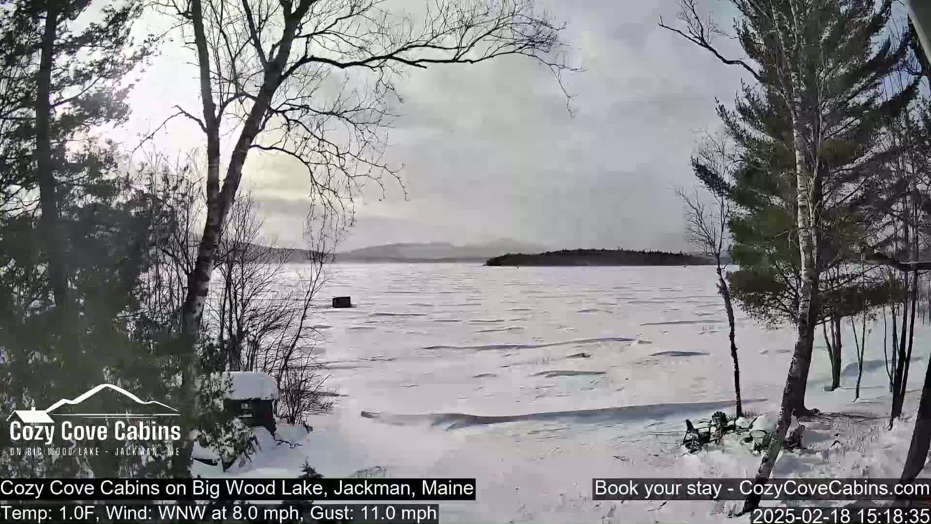 A partly cloudy, snowy winter scene shows a partially frozen lake with bare trees and a small cabin in the foreground.