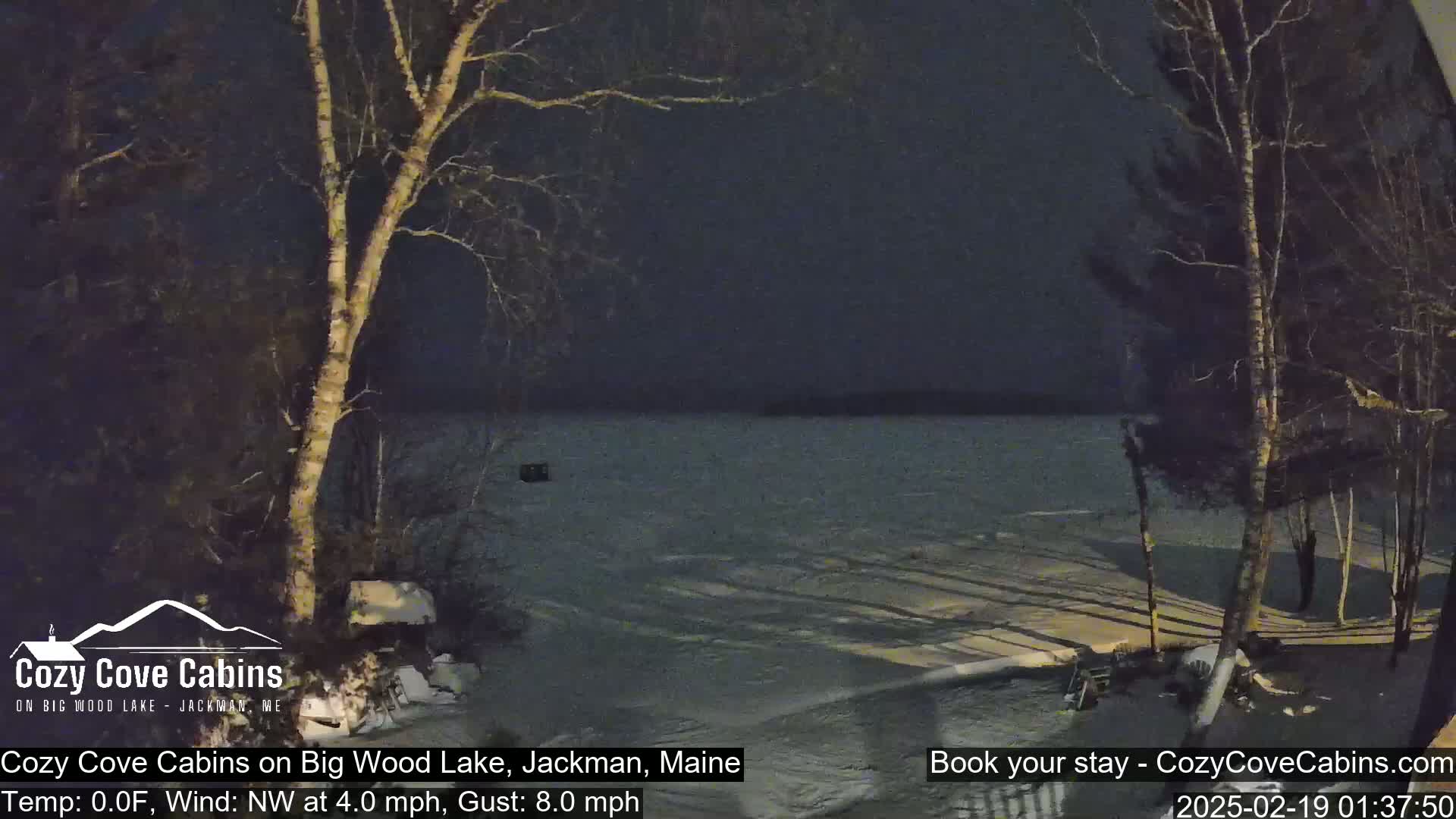 A nighttime view of a snow-covered lake shore with bare trees, under a dark sky with a northwest wind at 4 mph and gusts up to 8 mph.