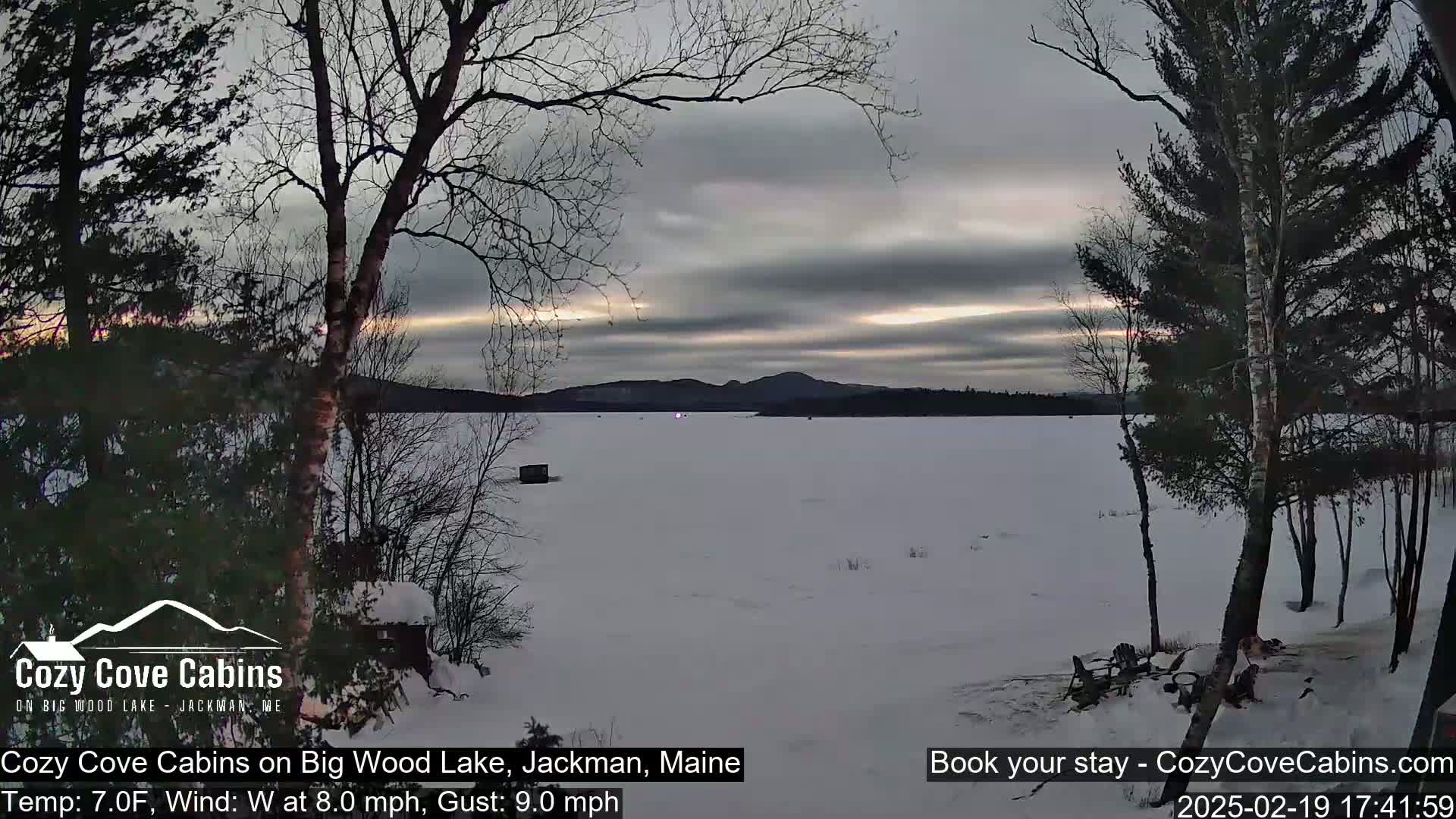 A snow-covered frozen lake is seen at dusk under a cloudy sky, with bare trees in the foreground and distant hills in the background.