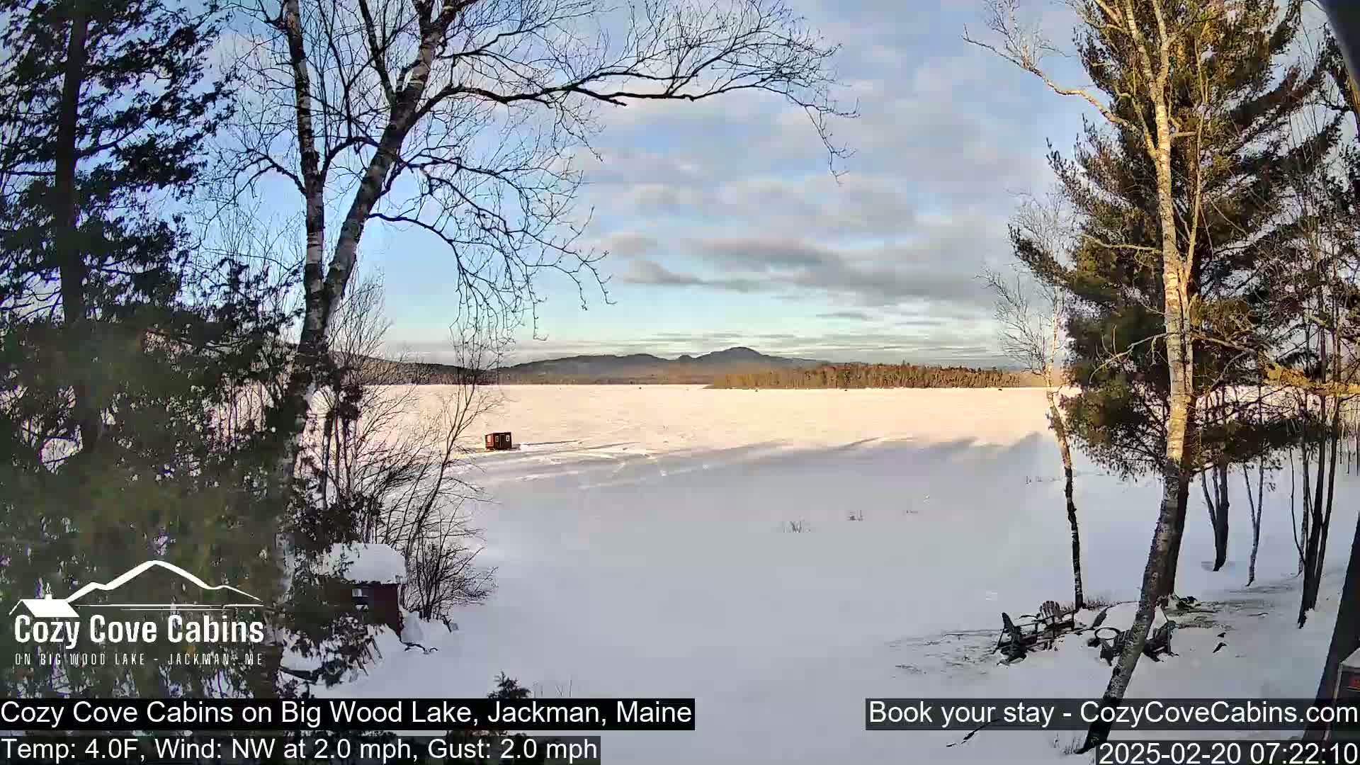 A snow-covered frozen lake is seen from a lakeside cabin, with a partly cloudy sky and bare trees in the foreground.