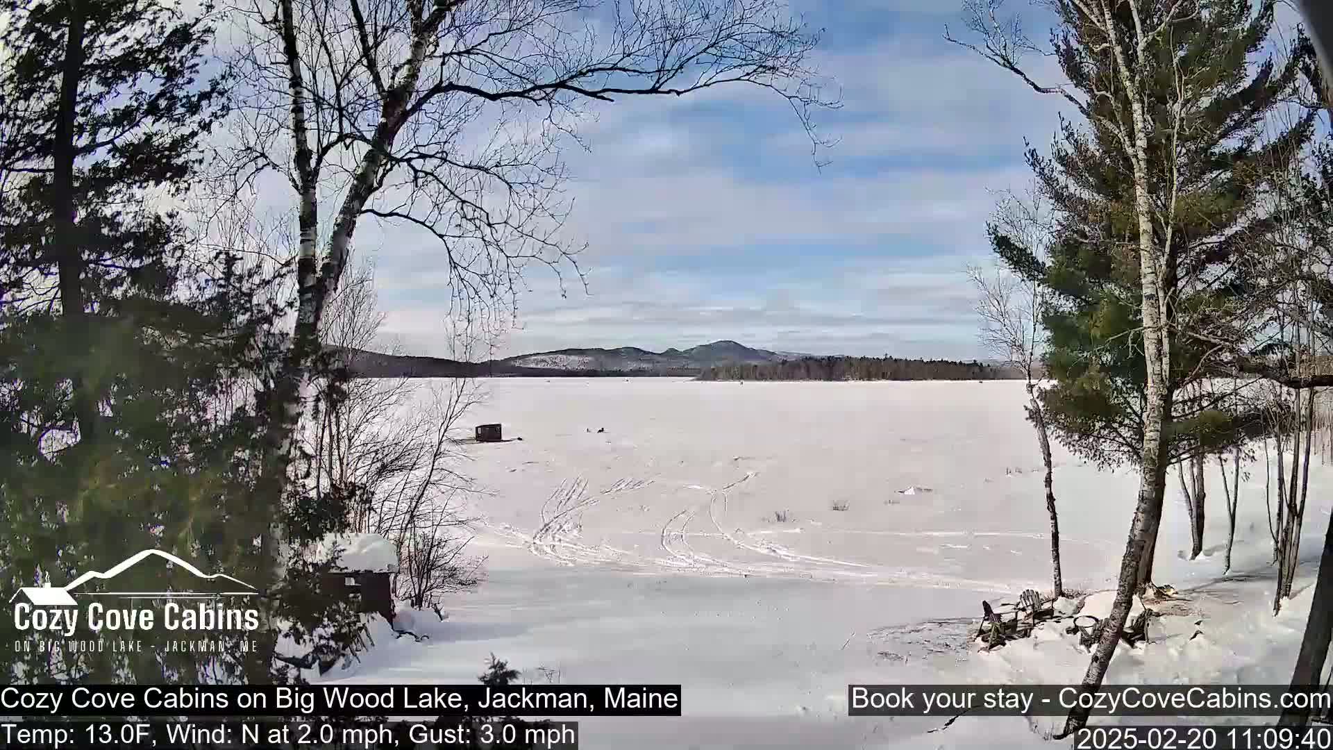 A snow-covered frozen lake stretches to distant, snow-capped mountains under a partly sunny sky, with bare trees in the foreground.