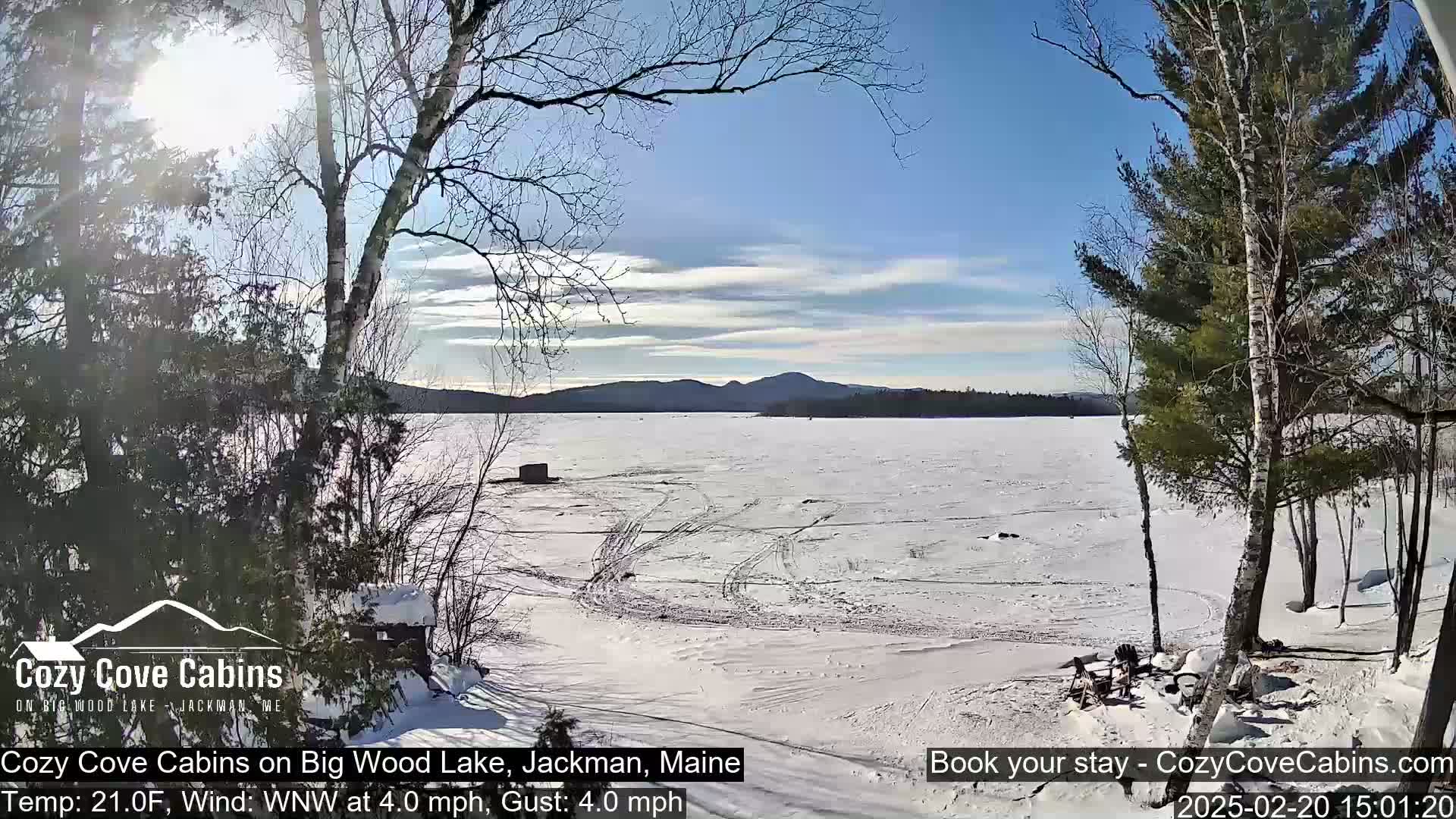 A frozen lake under a mostly sunny sky is viewed from a snow-covered shoreline with leafless trees and a small structure in the distance.