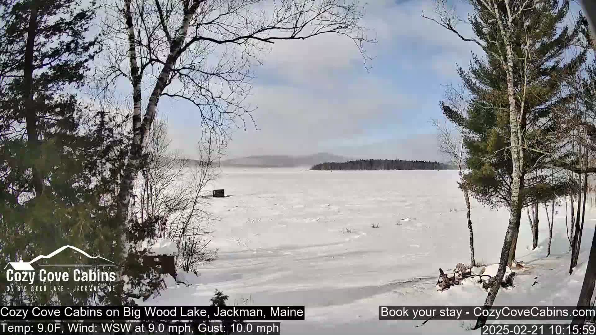 A snow-covered frozen lake is seen from a snowy shoreline, with leafless trees and a small structure visible in the foreground under a partly sunny sky with a light wind.