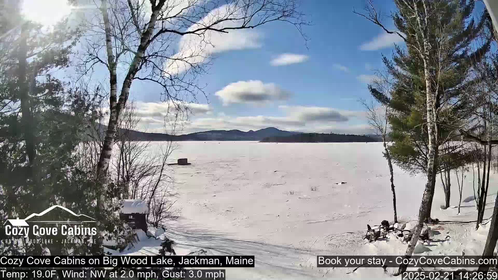 A snow-covered frozen lake is seen from a snowy shoreline, with bare trees and a small cabin in the foreground under a partly sunny sky.