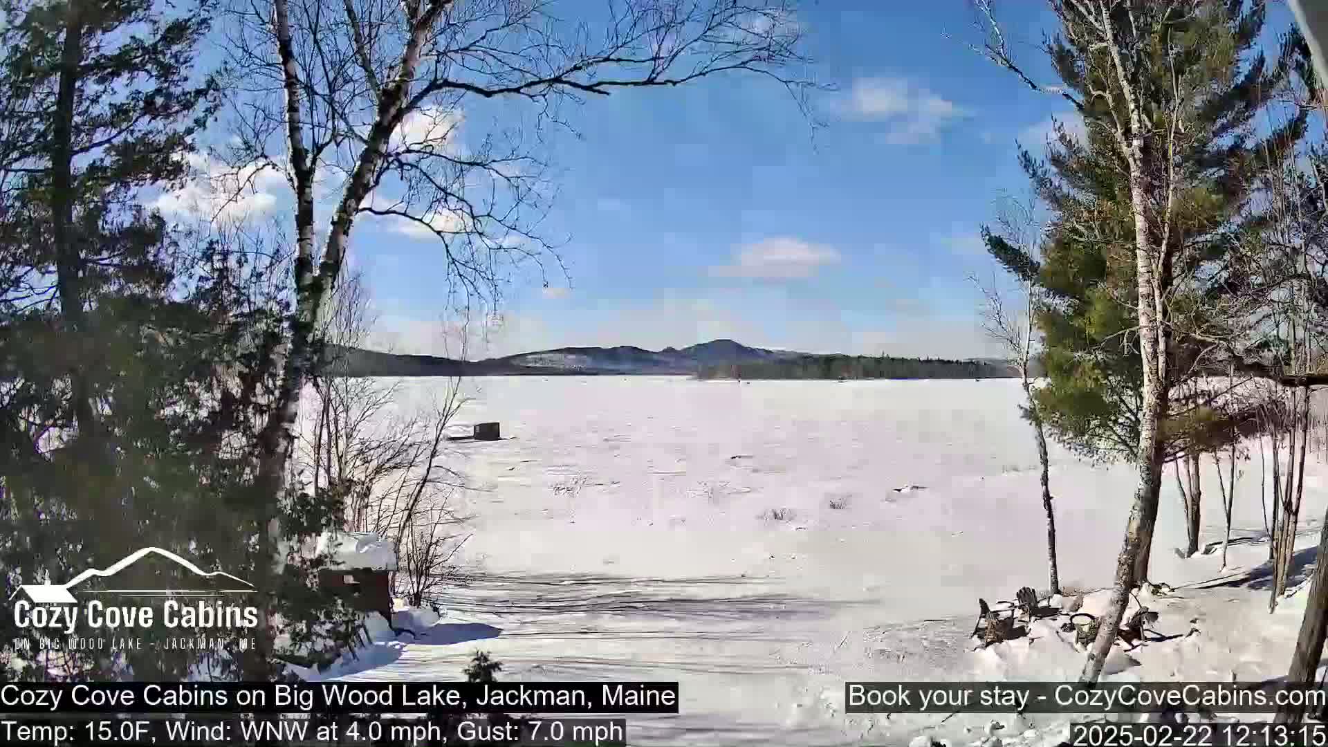 A snow-covered lake is seen from a snowy lakeside vantage point under a sunny sky with a few wispy clouds.