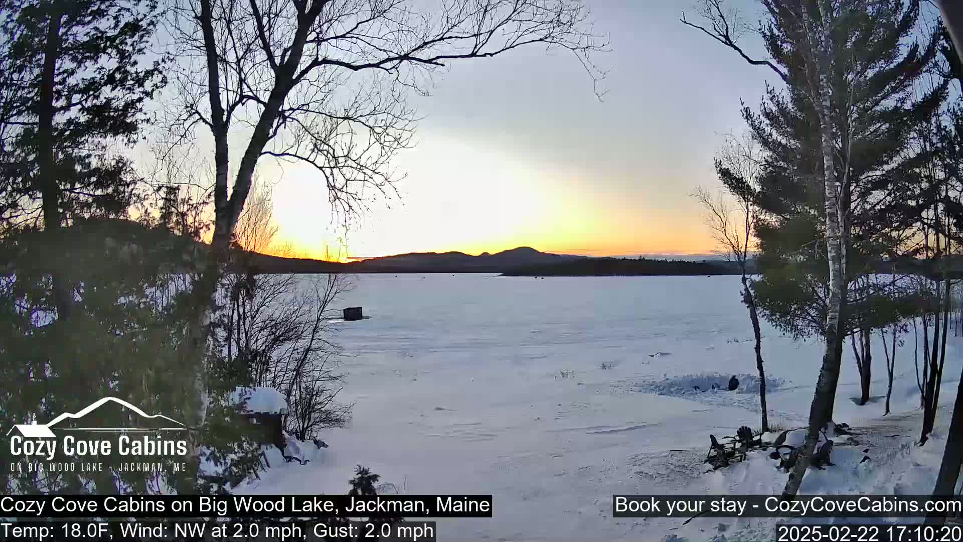 A frozen lake at sunset, under a mostly clear sky, is viewed from a snowy shoreline with bare trees.