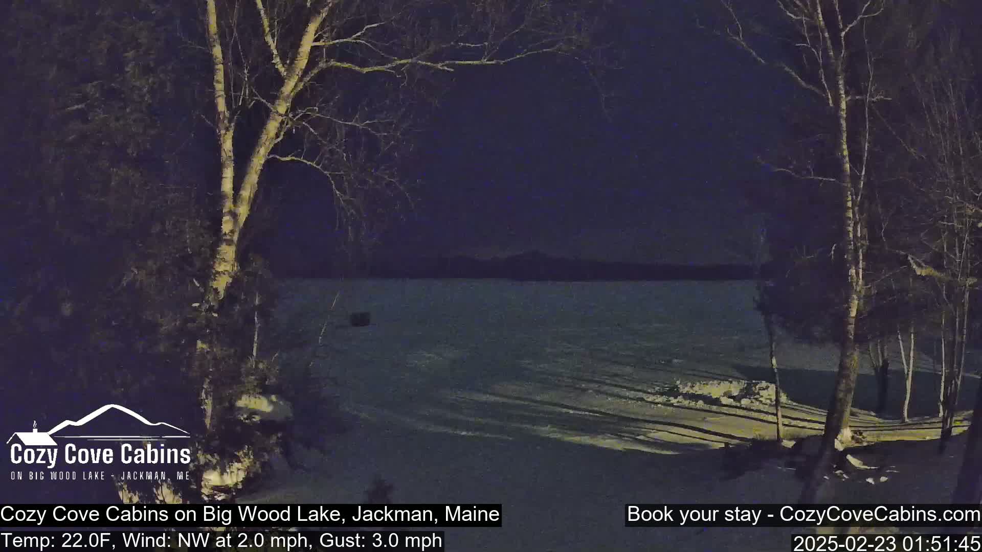 A nighttime view of a snow-covered lakeshore with bare trees under a dark sky, a slight northwest wind, and a temperature of 22 degrees Fahrenheit.
