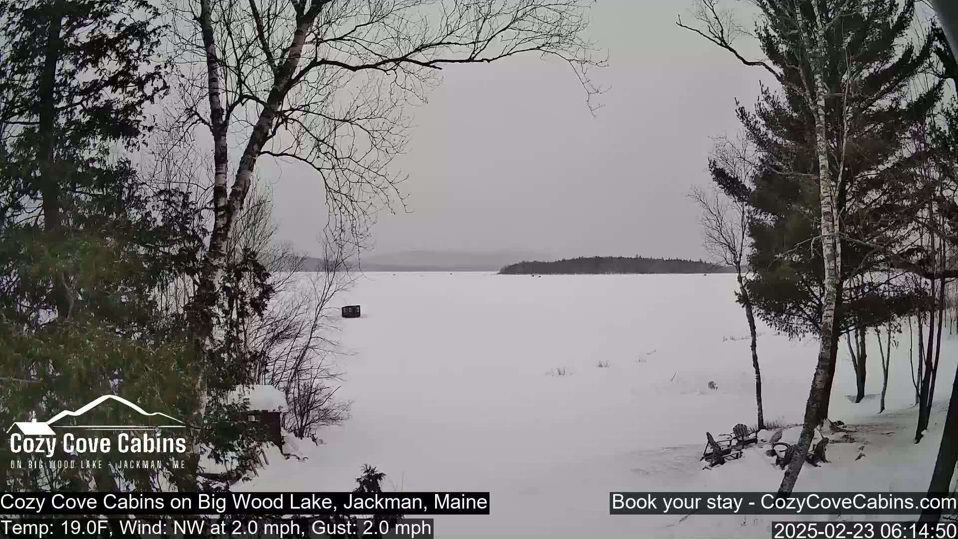 A snow-covered frozen lake is seen from a snowy shoreline with leafless trees and a small structure in the distance under an overcast sky.
