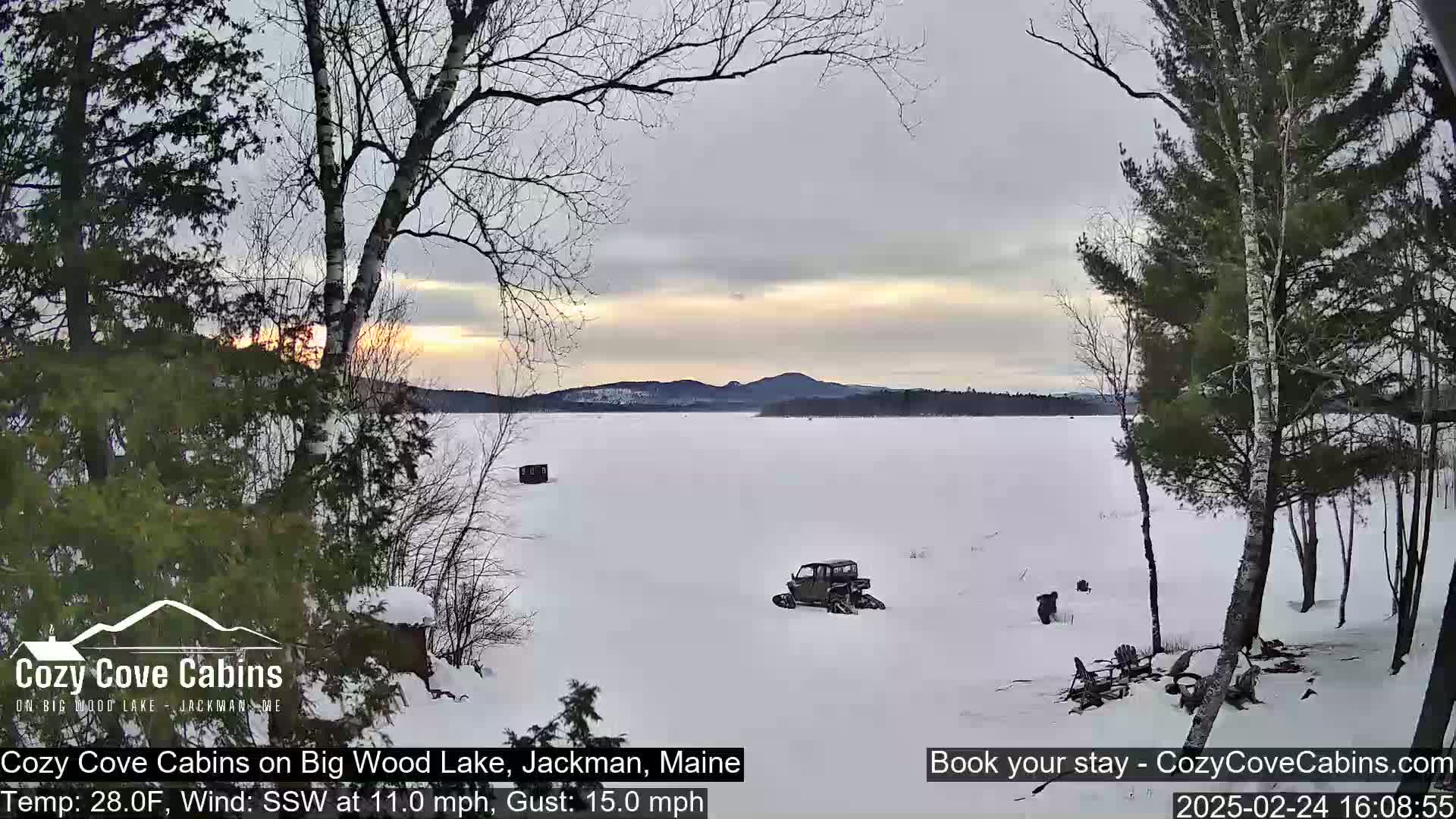 A snow-covered frozen lake at sunset is viewed from a snowy shoreline with trees, a utility vehicle, and some other objects on the ice.