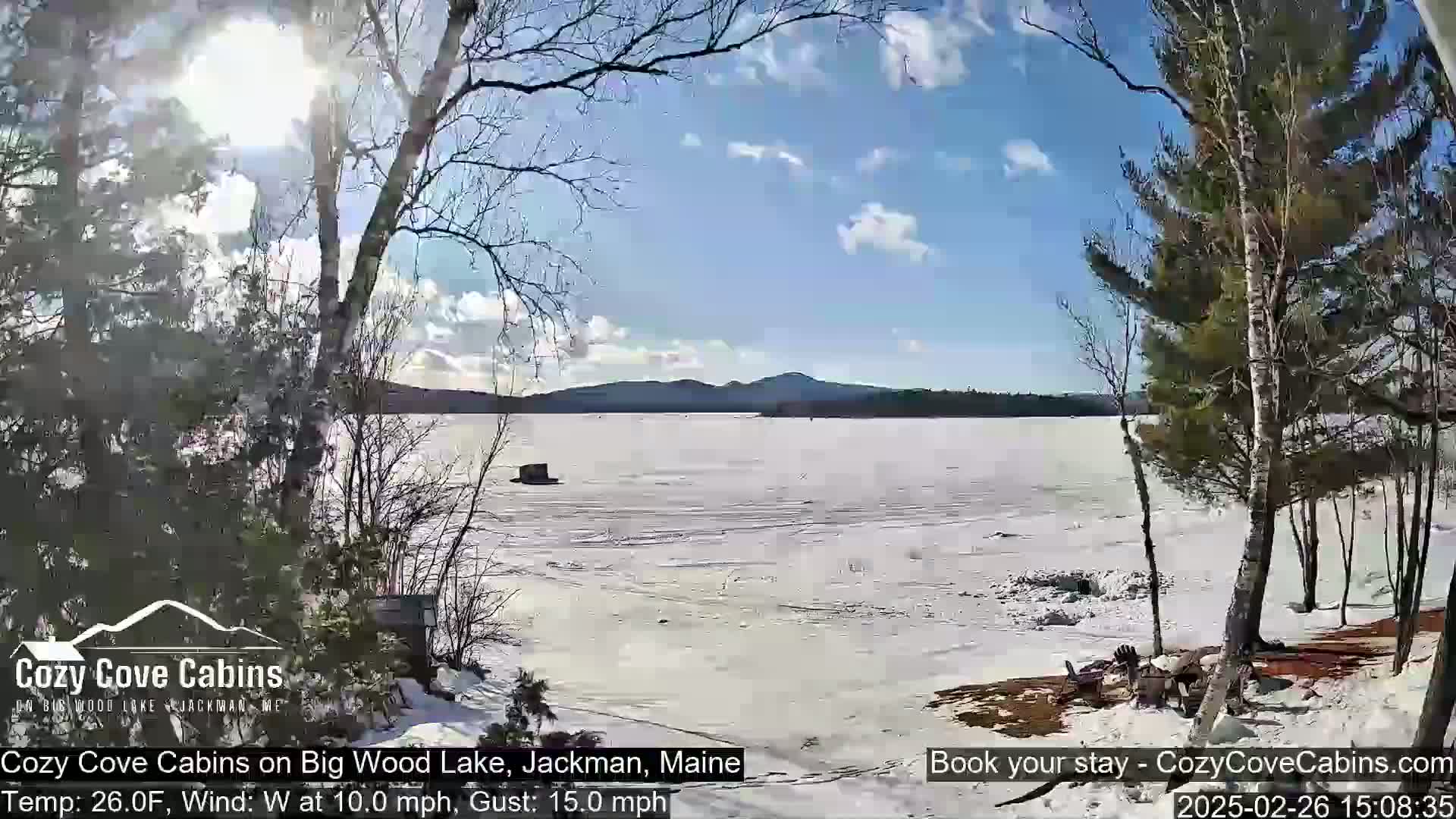 A partly sunny day with a westerly wind shows a mostly frozen lake with distant mountains under a blue sky, viewed from a snow-covered shoreline with bare trees.
