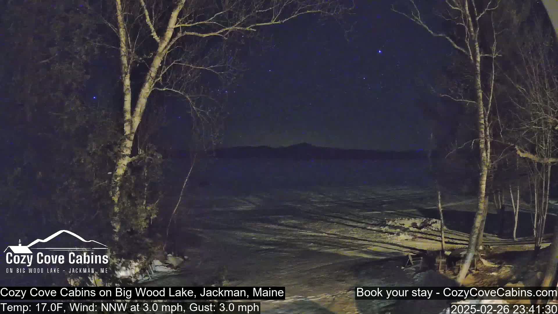 A night scene of a snow-covered lake under a starry sky, with bare trees in the foreground and a slight northwest wind.