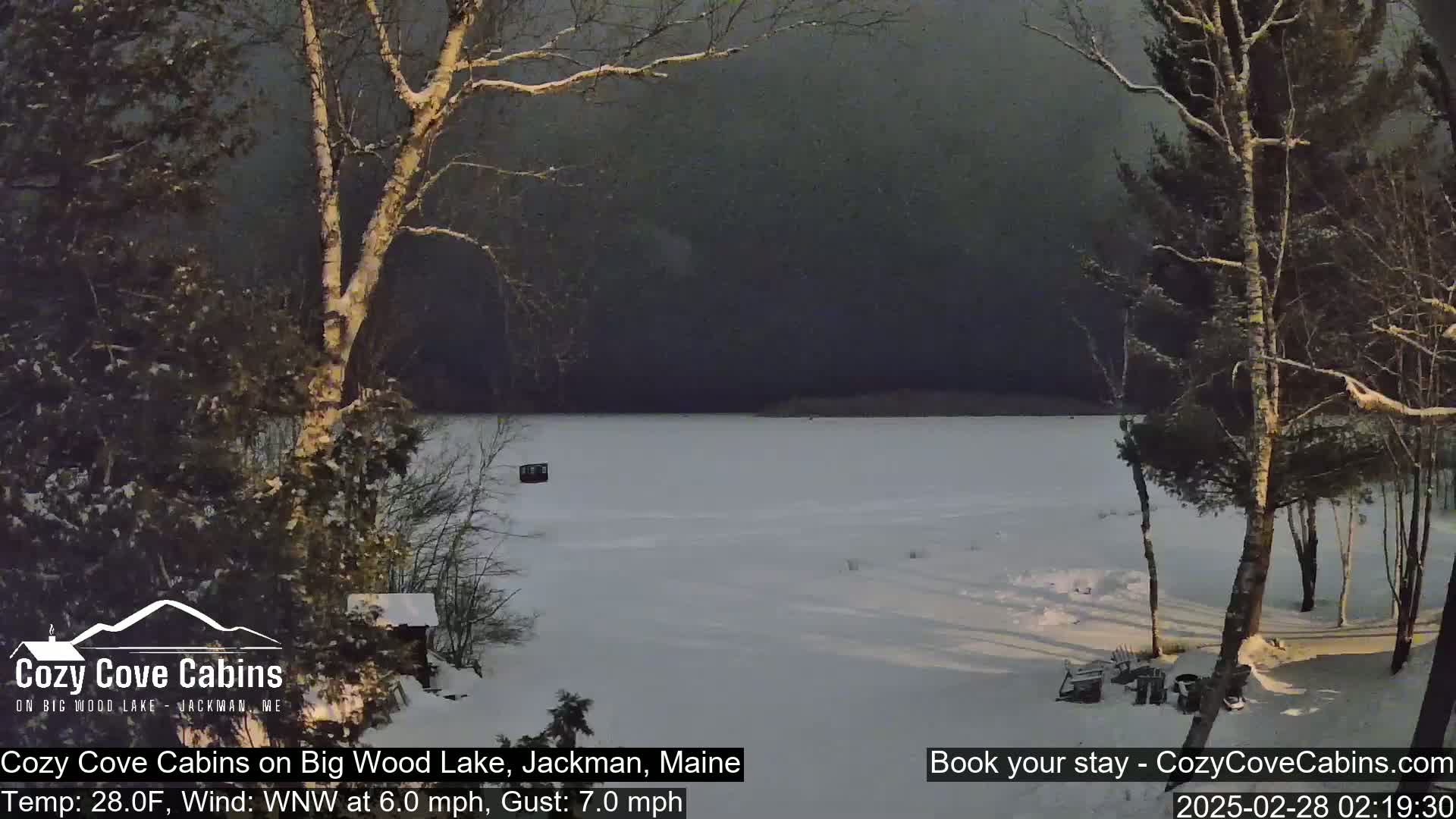 A frozen lake is seen from a snowy shoreline, under a dark, overcast sky.