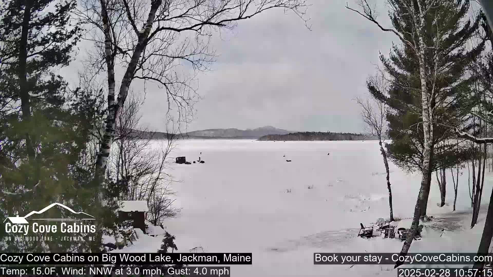 A snow-covered frozen lake is viewed from a snowy shoreline, with bare trees and a small cabin visible in the foreground under an overcast sky.