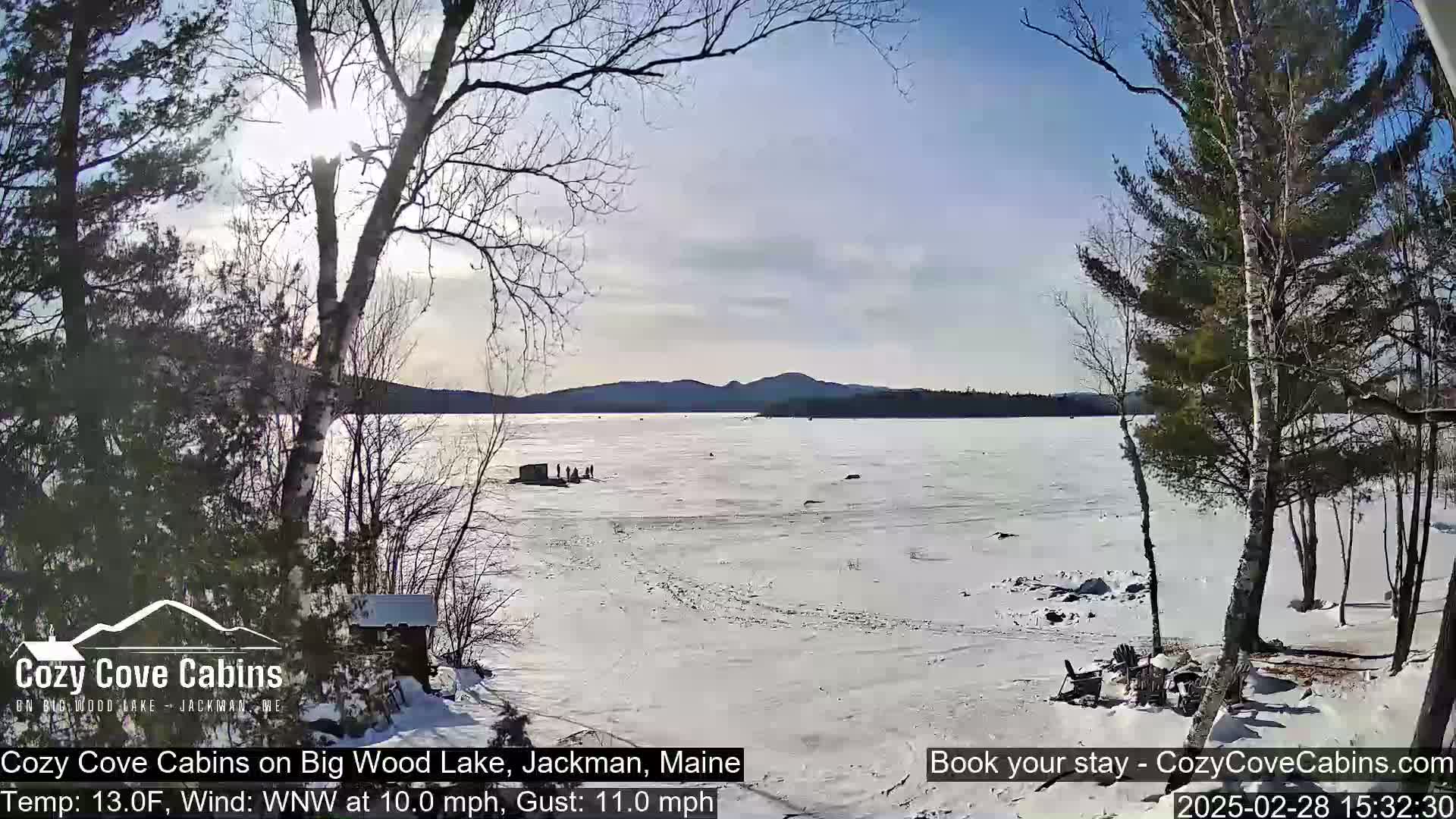A sunny, cold day on a snow-covered frozen lake with distant mountains under a partly cloudy sky, viewed from a snow-covered shoreline with leafless trees.