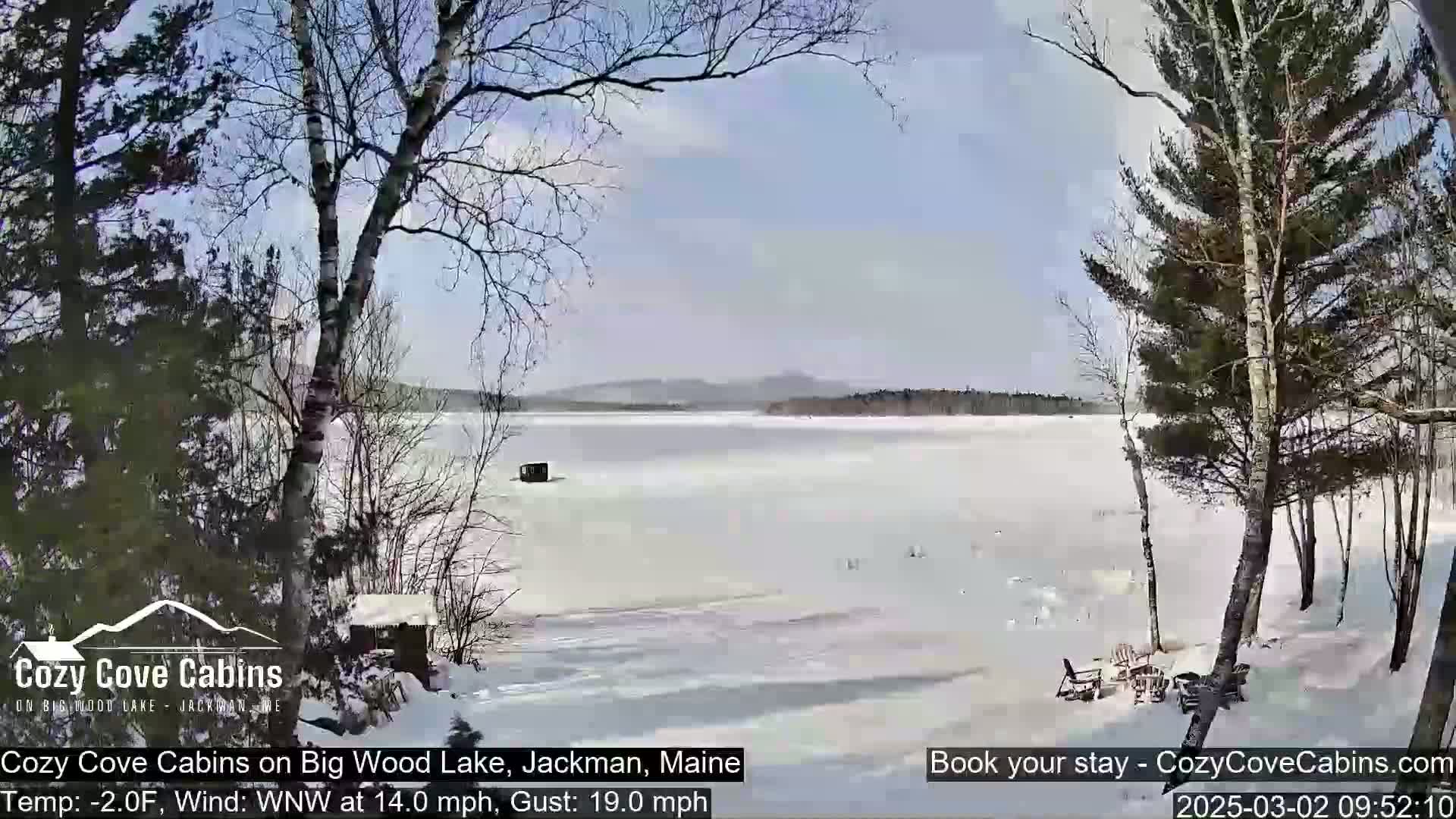 A mostly sunny, cold day on a snow-covered frozen lake is seen from a snowy shoreline, with leafless trees and a small structure visible in the distance.