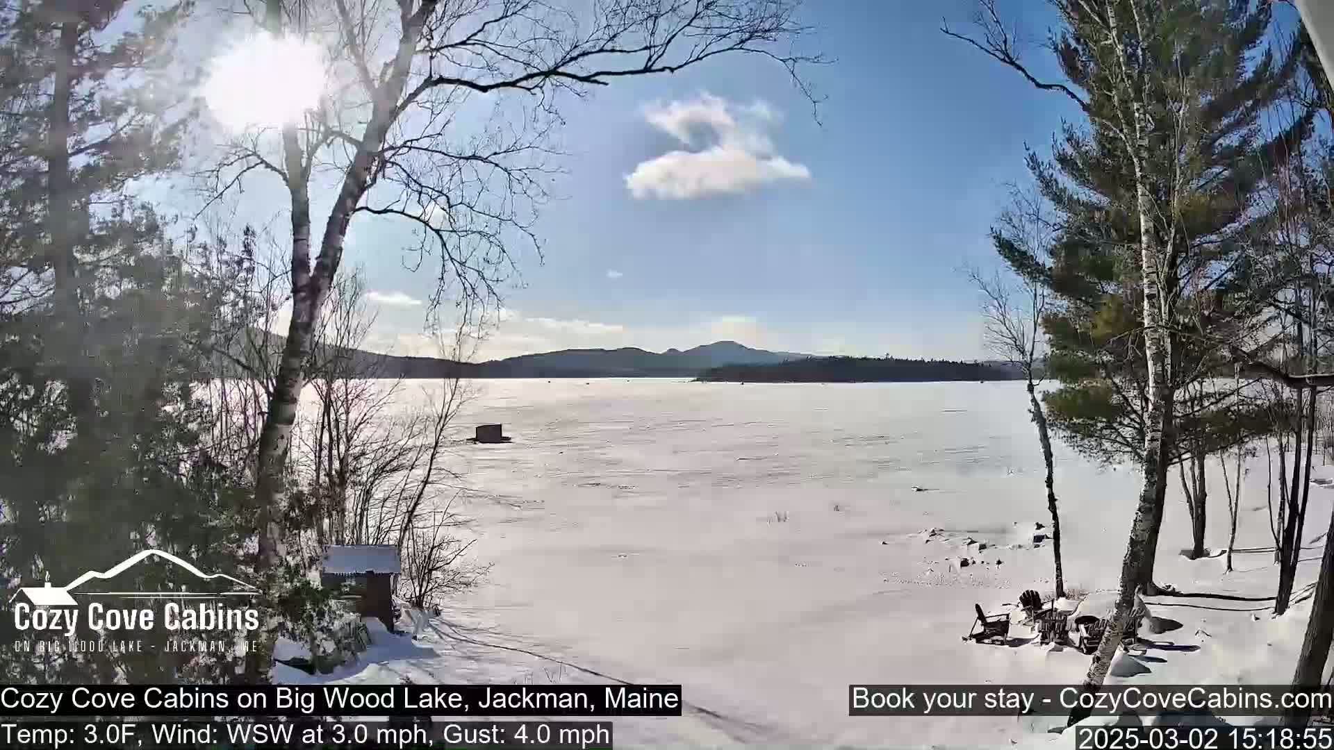 A sunny, snowy scene shows a frozen lake with distant hills under a partly cloudy sky, viewed from a snow-covered lakeshore with leafless trees.
