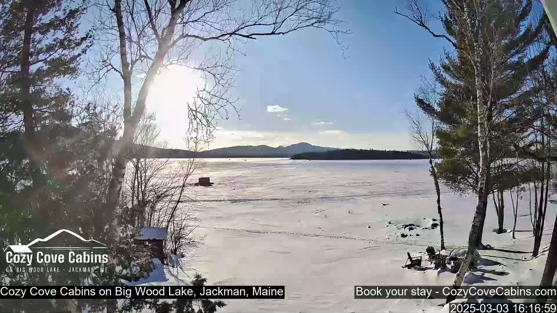A sunny winter day shows a snow-covered frozen lake with distant mountains under a mostly clear sky, viewed from a snowy shoreline with leafless trees.