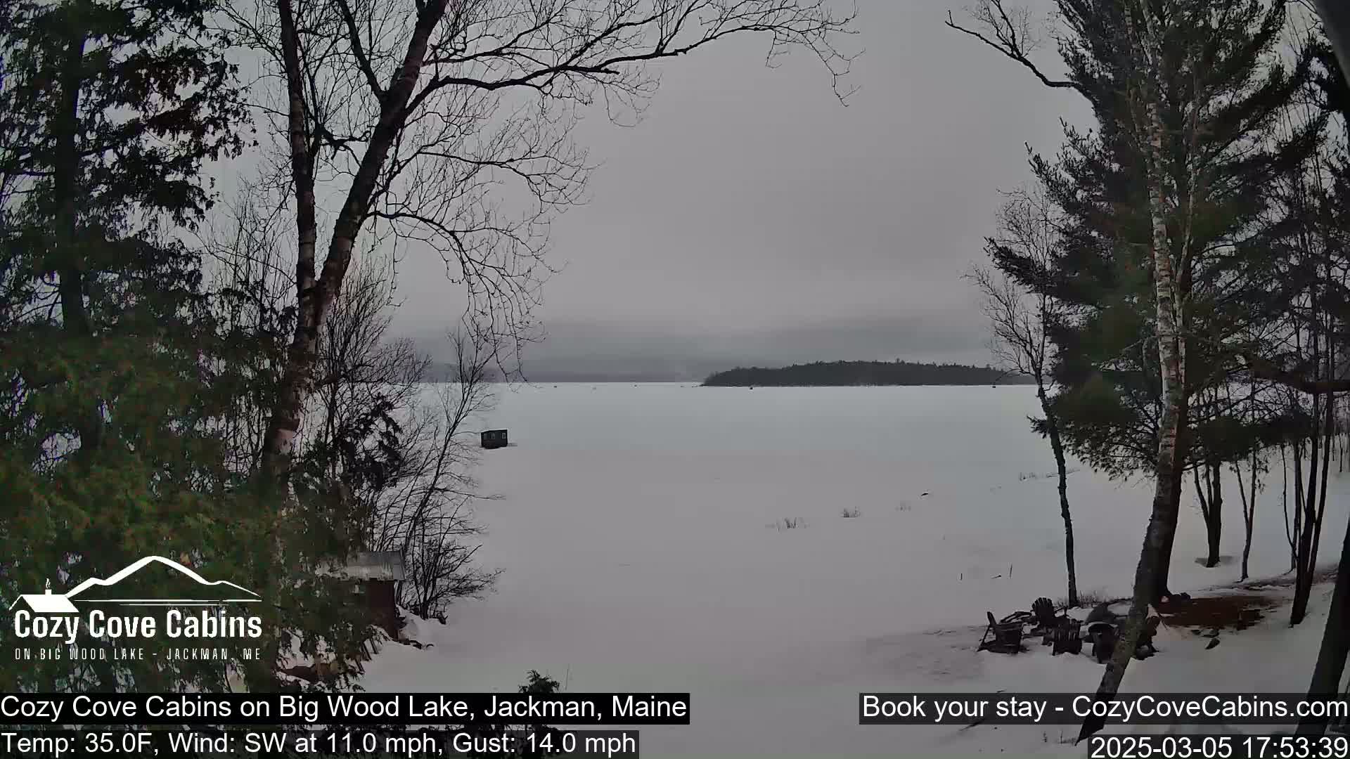 A snow-covered frozen lake is seen from a snowy shoreline, under an overcast sky, with bare trees in the foreground and a small structure on the ice in the mid-ground.