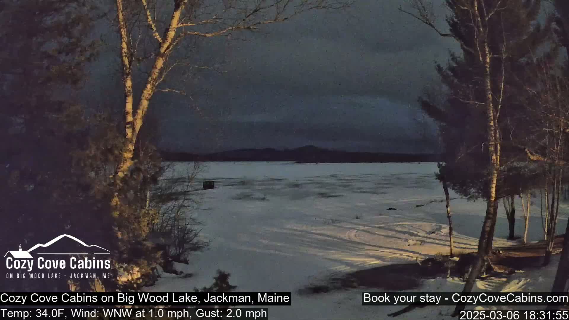 A snow-covered frozen lake is seen at dusk, under a dark, cloudy sky, with bare trees in the foreground.