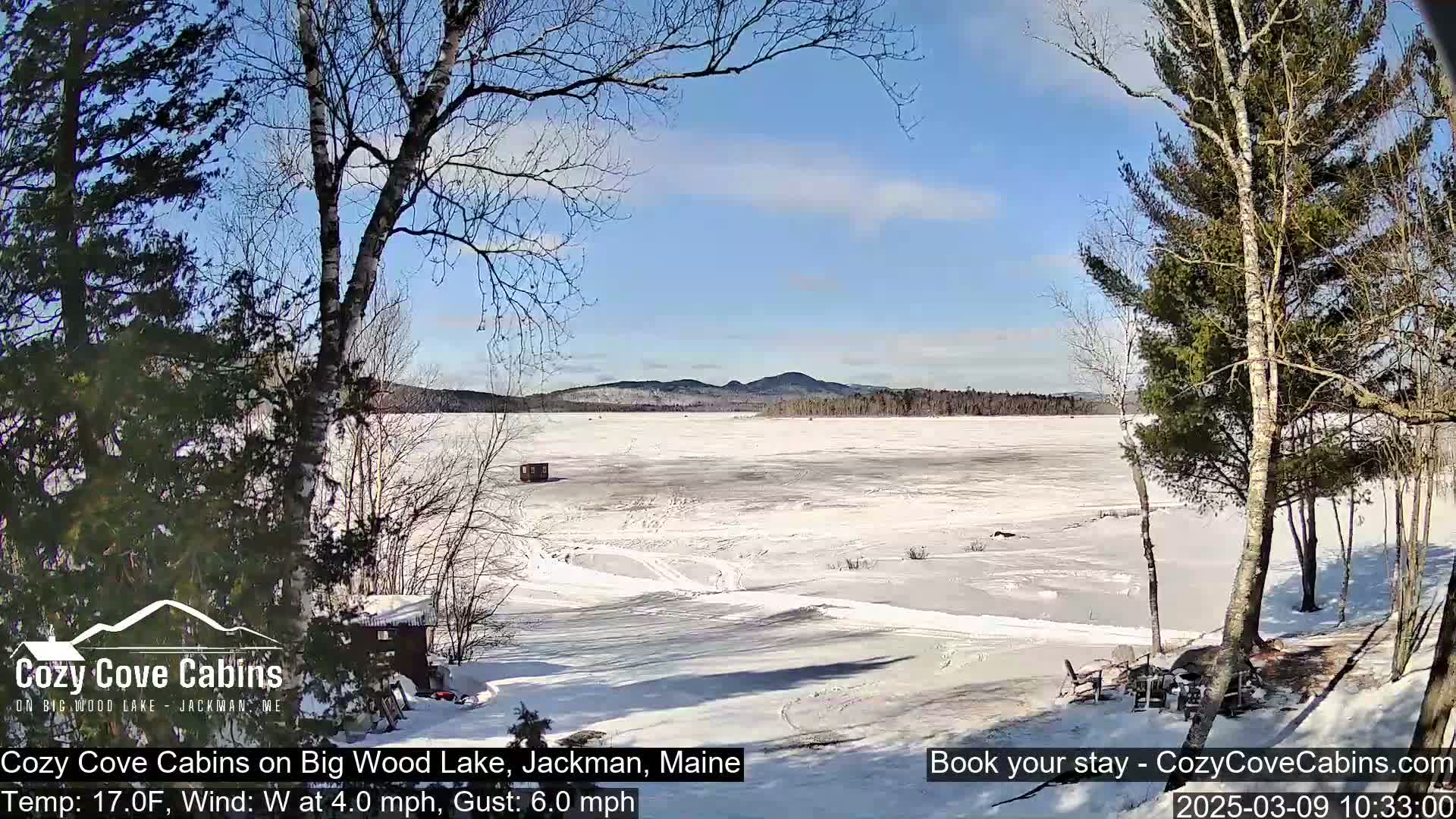 A snow-covered frozen lake is seen from a wooded shoreline on a sunny day with a light westerly wind.