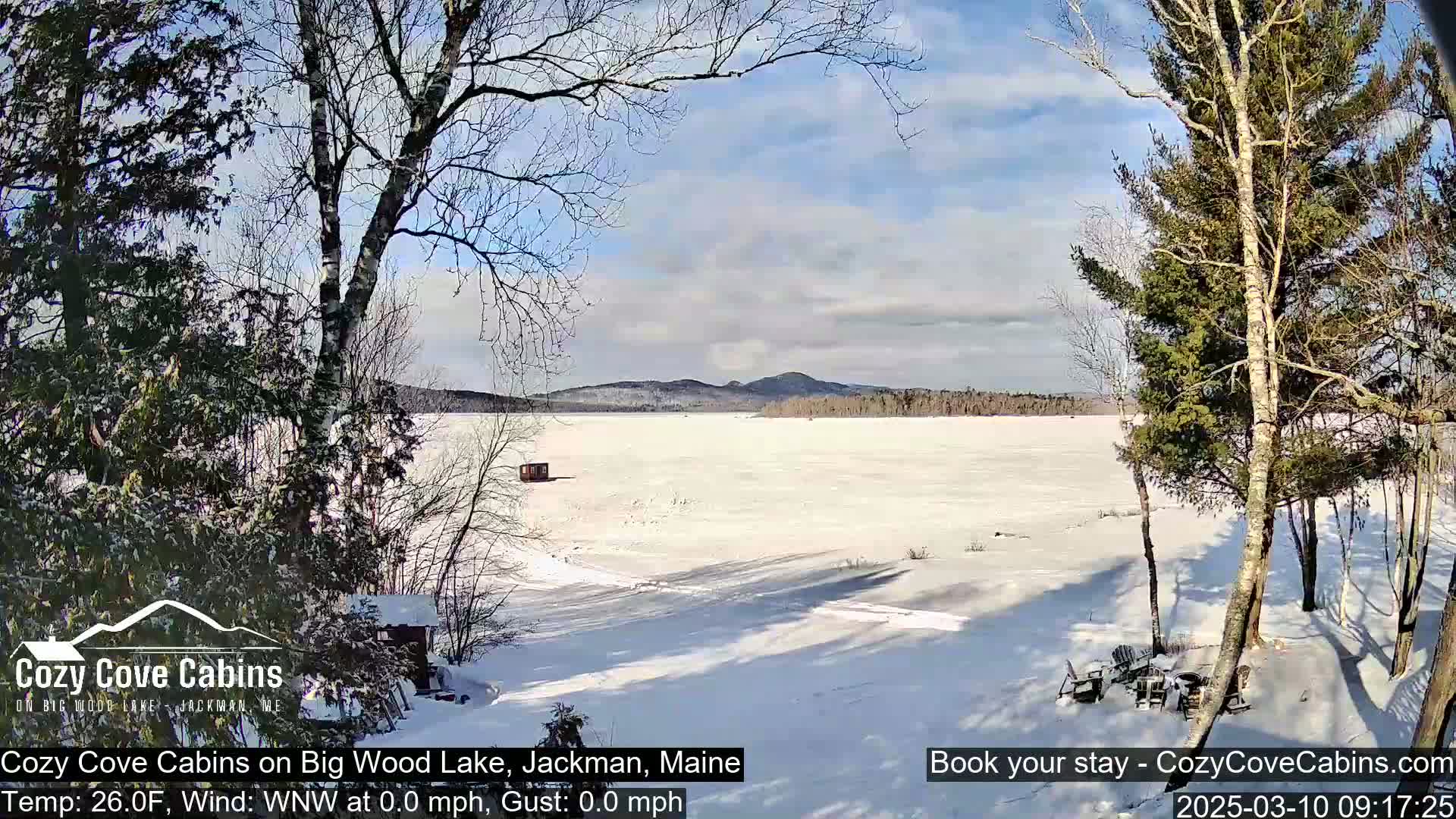 A snow-covered frozen lake is seen from a snowy shoreline, with bare trees and a few evergreen trees in the foreground under a partly cloudy, sunny sky.
