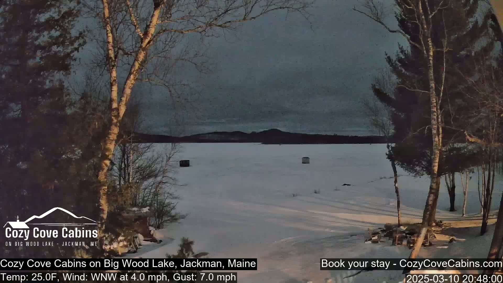 A snow-covered frozen lake is visible at dusk, under a gray sky, with a few ice fishing shelters on the ice and bare trees in the foreground.