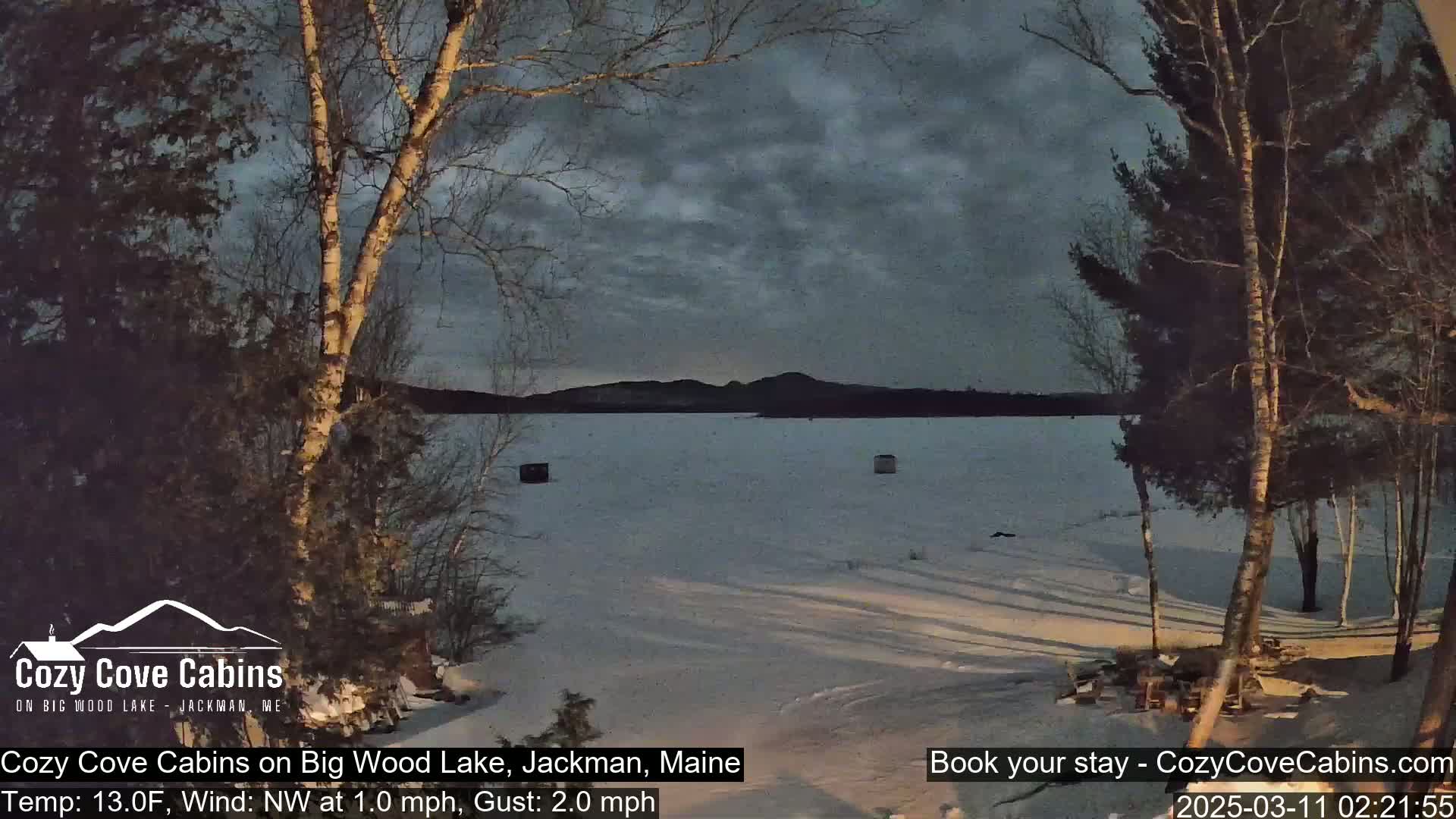 A frozen lake is seen at dusk under a cloudy sky, framed by snow-covered trees.