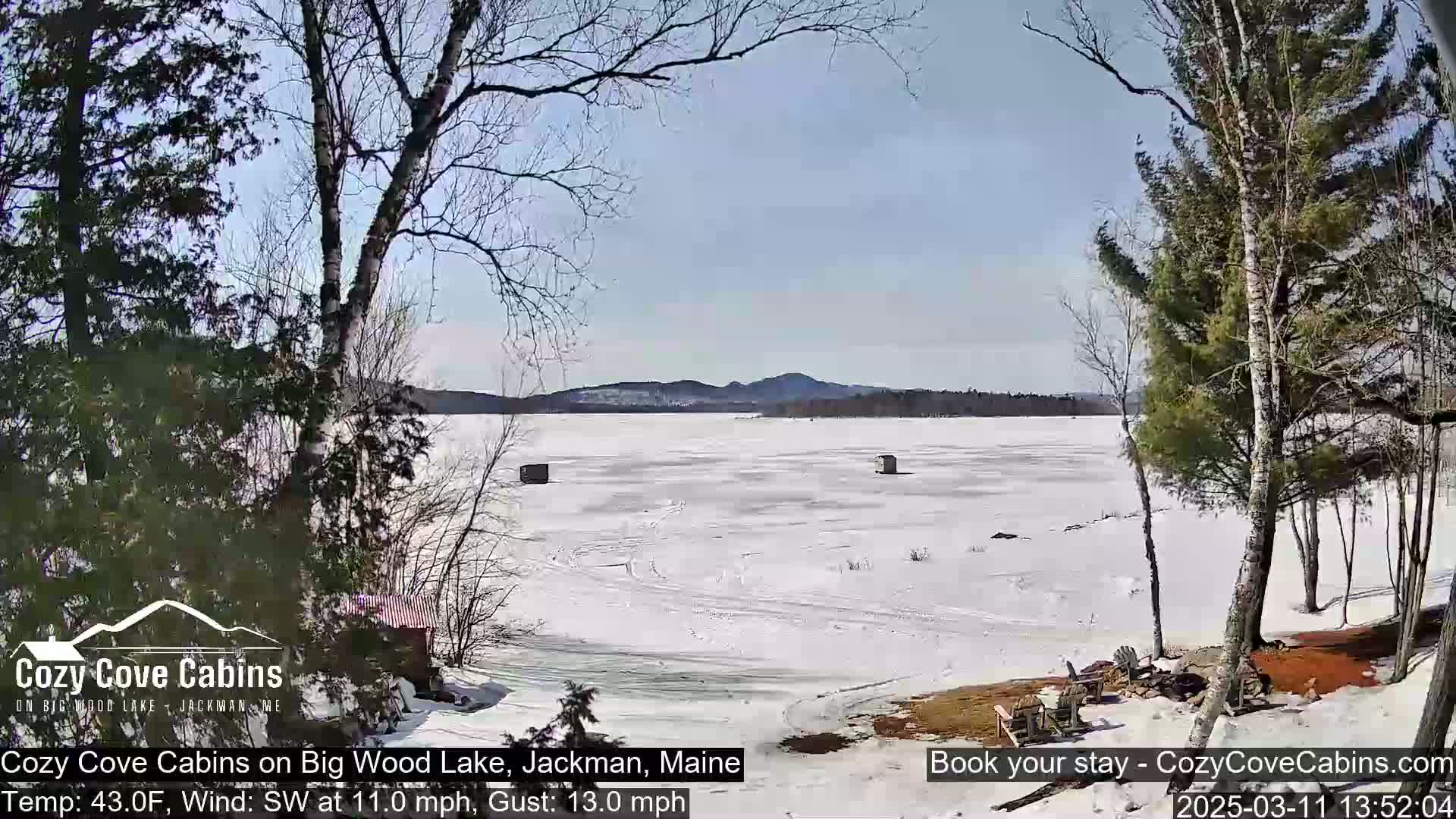 A snow-covered, frozen lake is seen from a lakeside vantage point under a partly sunny sky, with bare trees and two small structures visible on the ice.