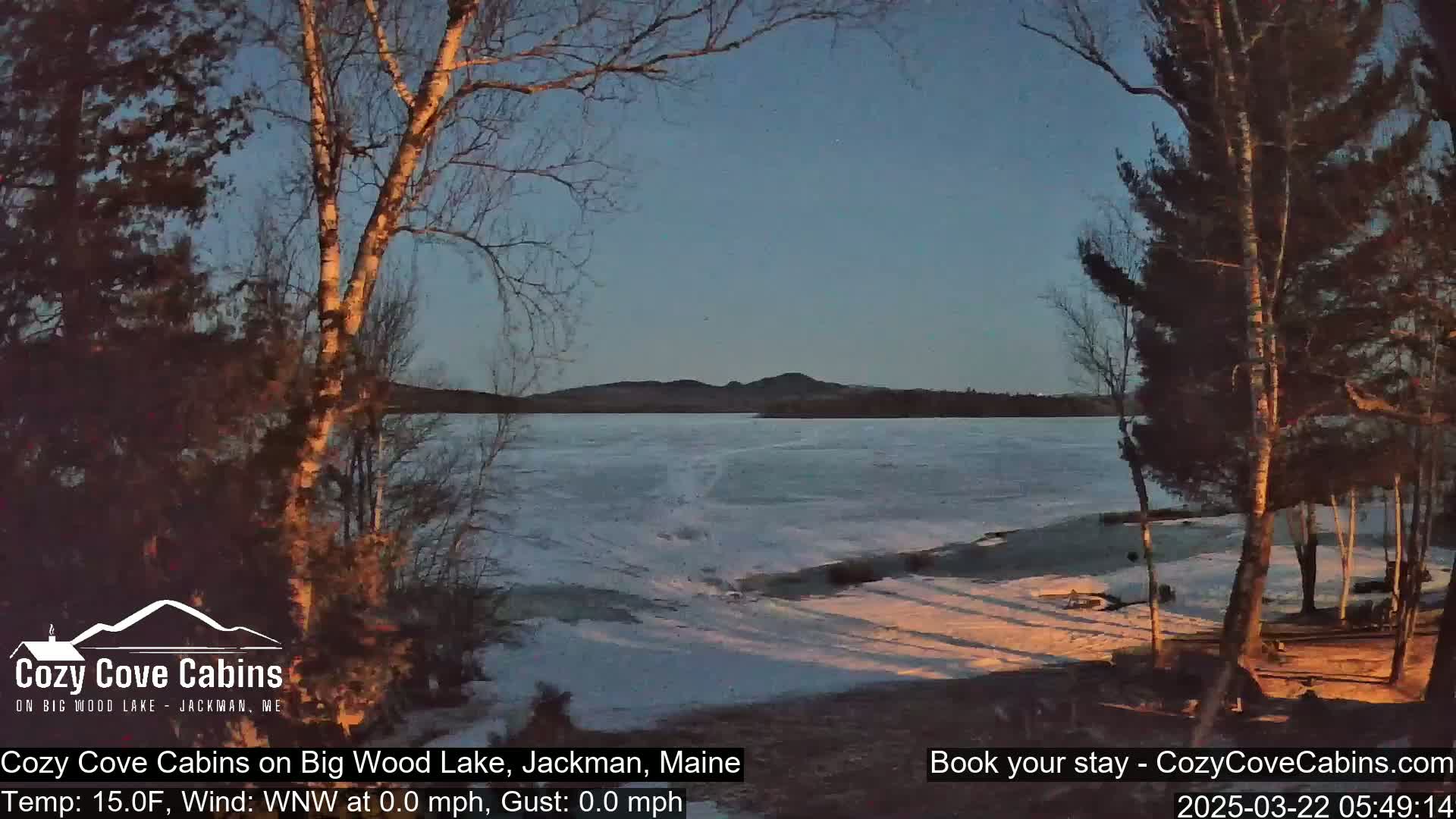 A partially frozen lake at sunset is viewed from a snow-covered shoreline, with bare trees in the foreground and a calm, clear sky.