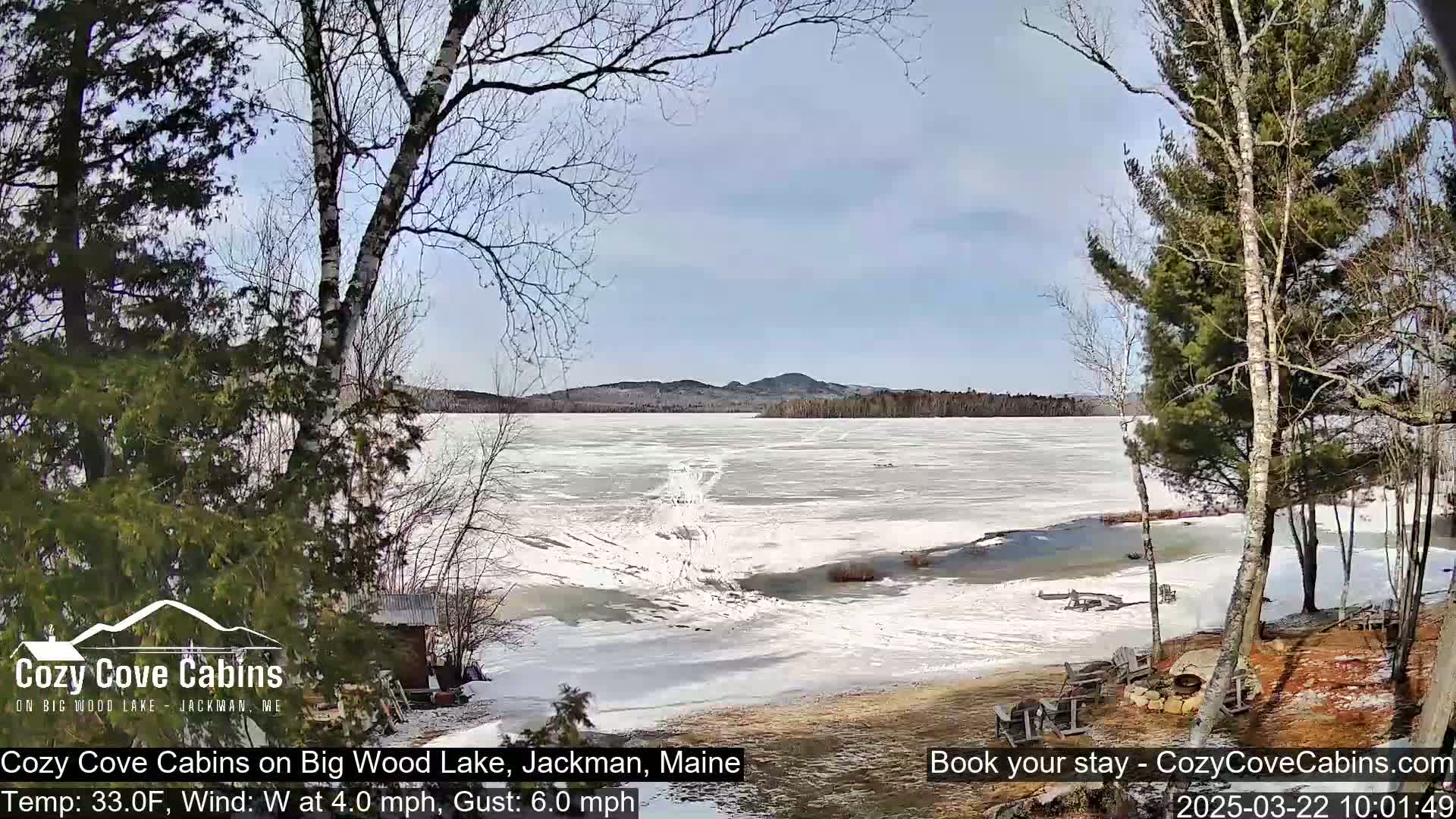 A partially frozen lake is viewed from a snow-covered shoreline, under a partly sunny sky, with leafless trees in the foreground.