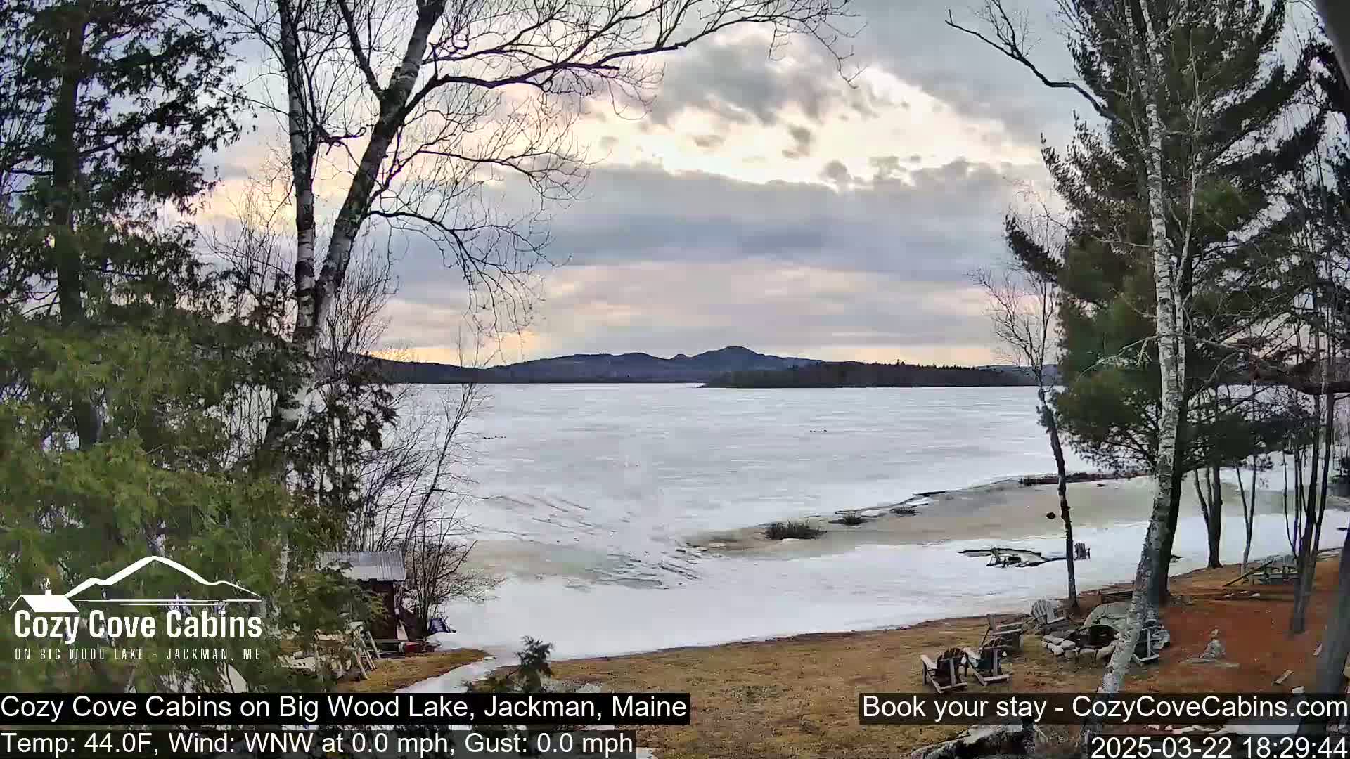 A partially frozen lake is seen from a lakeside yard with bare trees under a mostly cloudy sky.