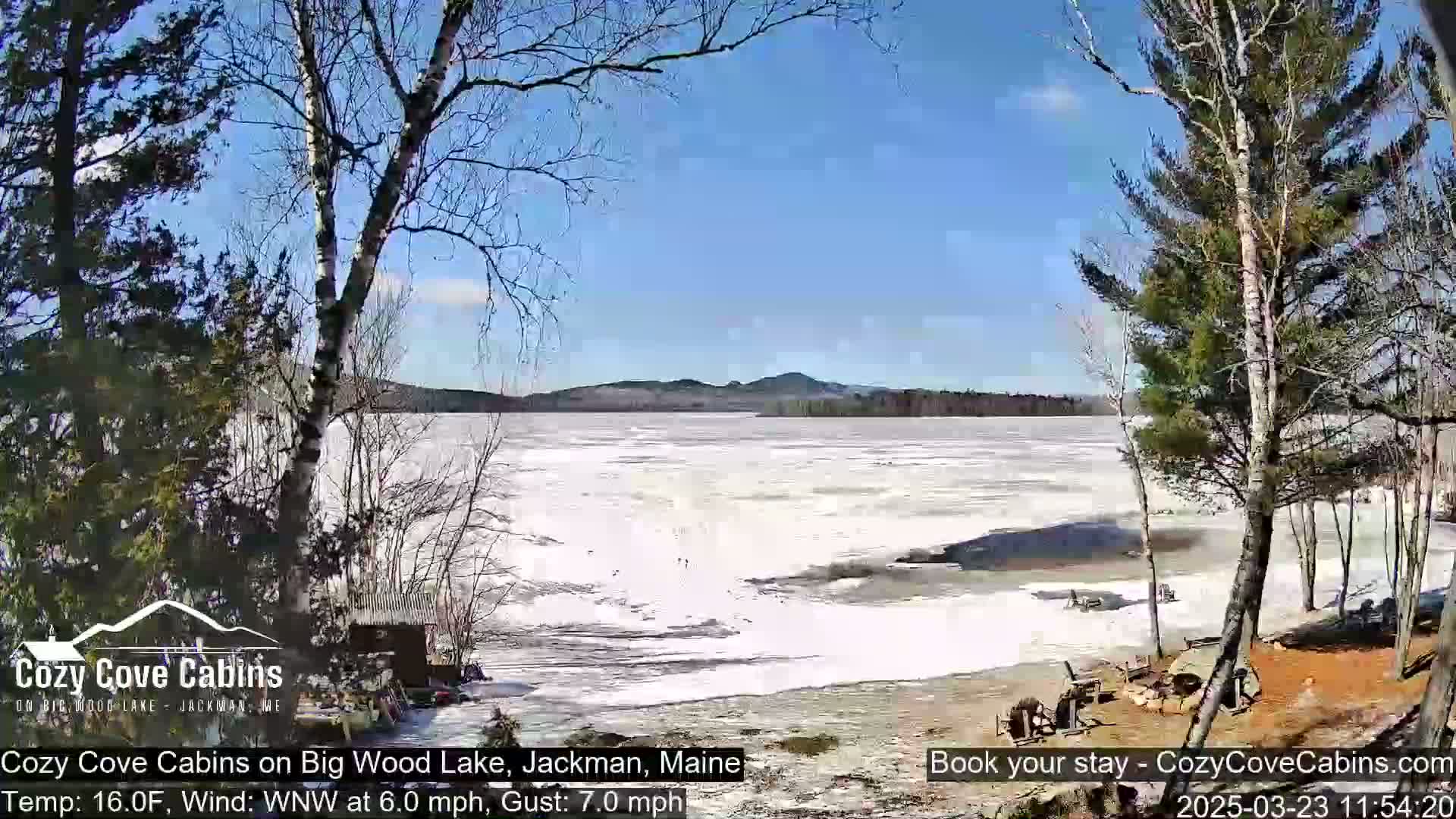 A partially frozen lake is seen from a lakeside vantage point on a sunny day with a light wind.